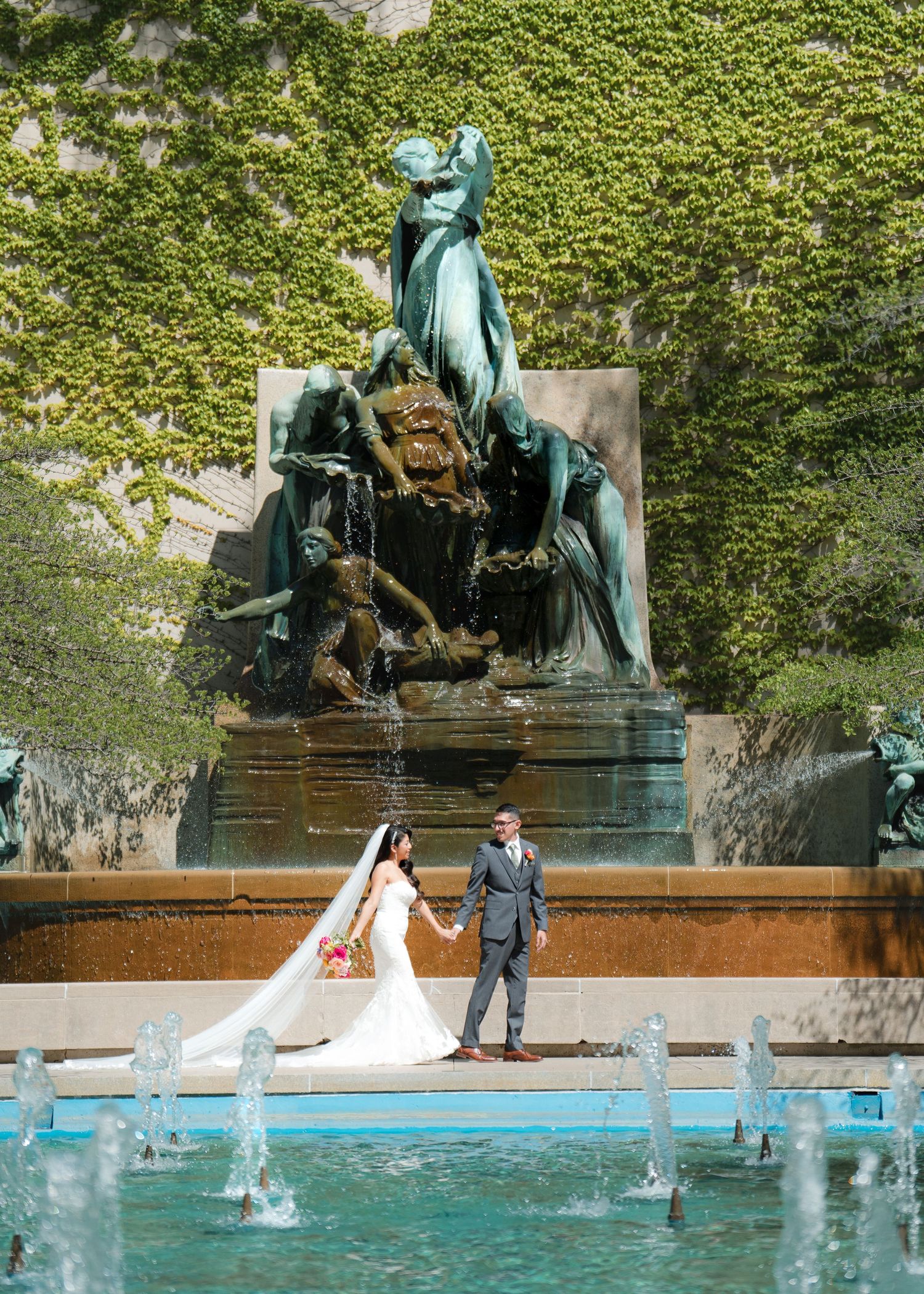 Bride and groom hold hands before ornate bronze fountain sculpture with ivy-covered wall backdrop on wedding day.