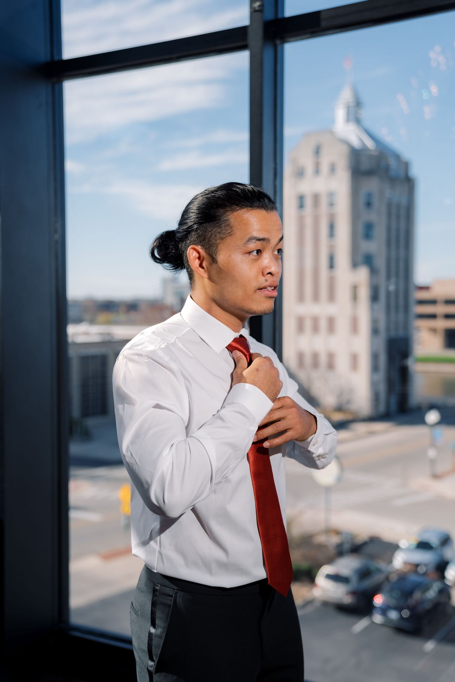 Person adjusting red tie while standing near window with city view.