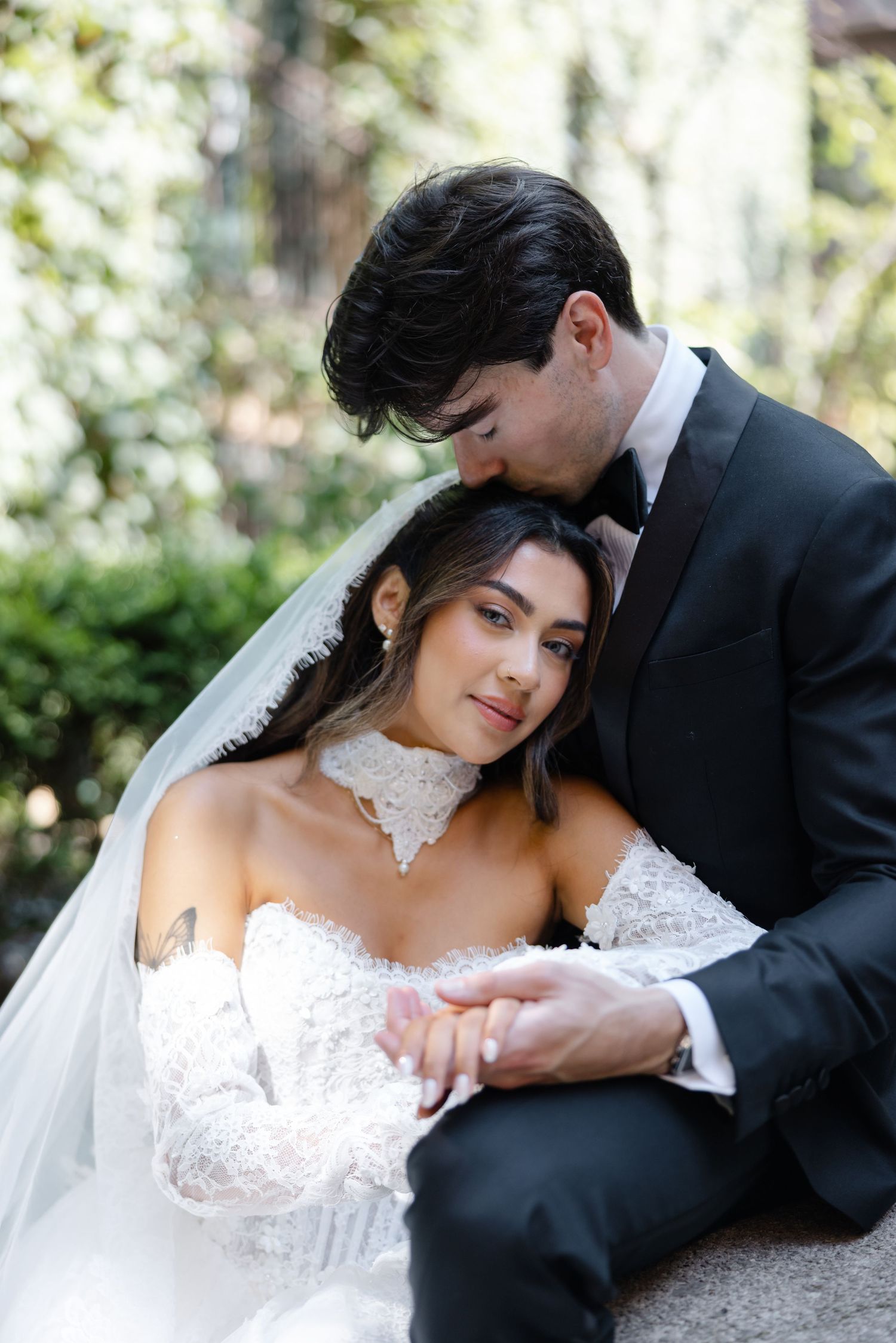 A groom in a black tuxedo kisses his bride wearing a lace off-shoulder gown and veil outdoors.