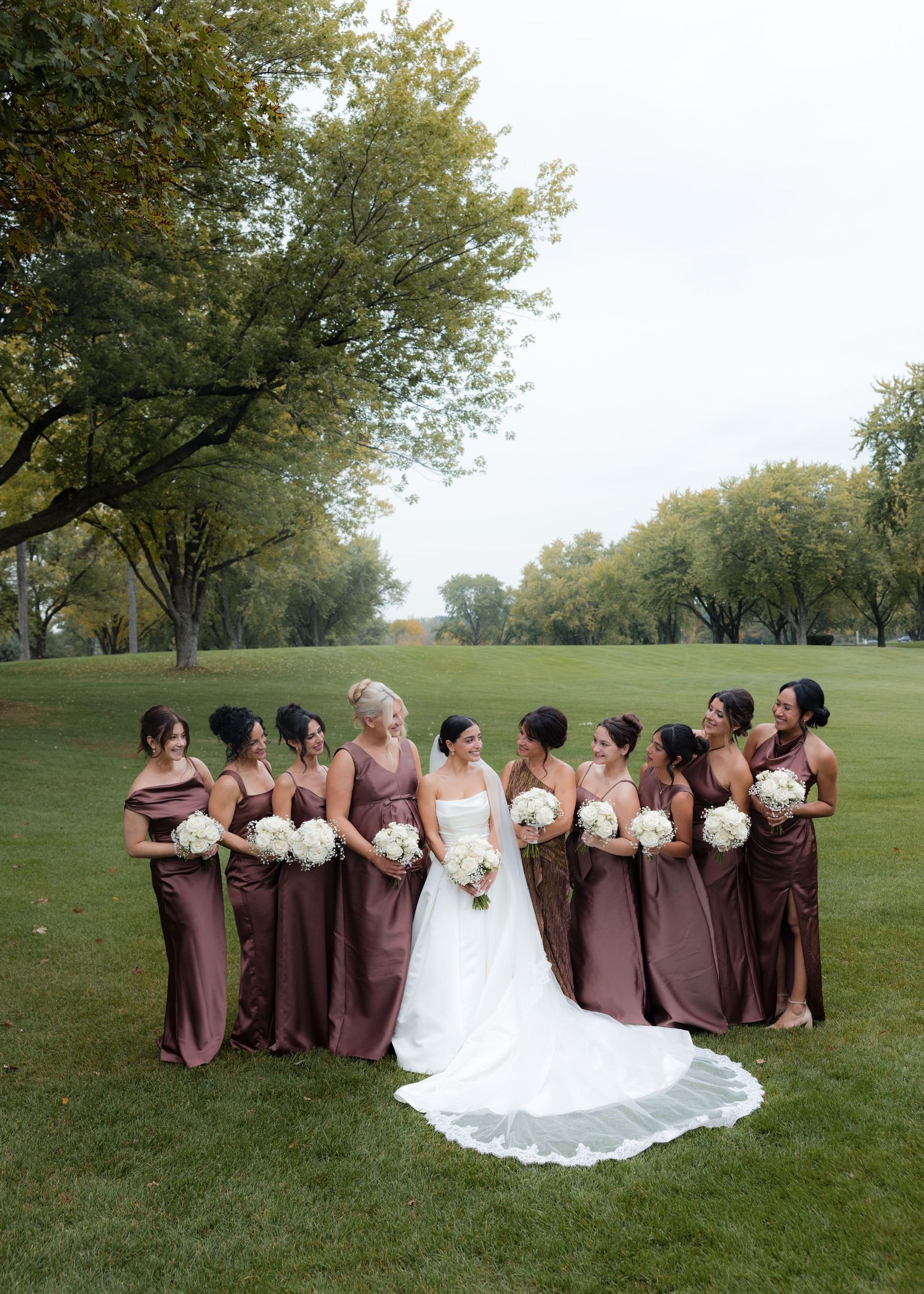 Wedding party in brown dresses poses on a green lawn with trees in the background.