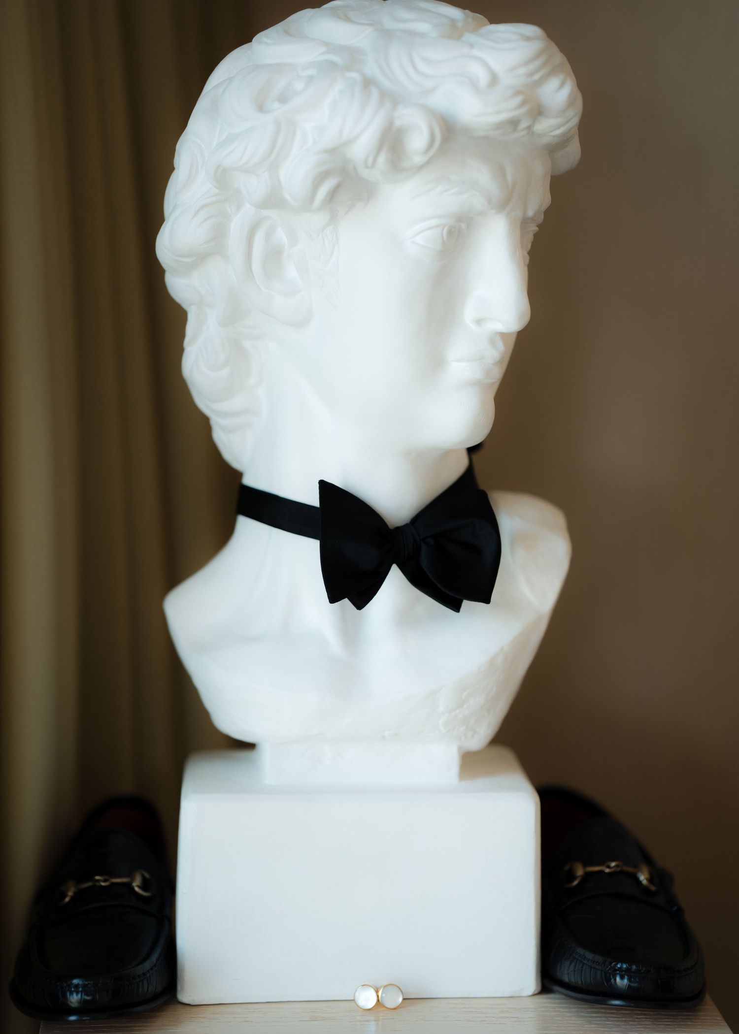 Classical white bust sculpture wearing black bowtie displayed with formal shoes.