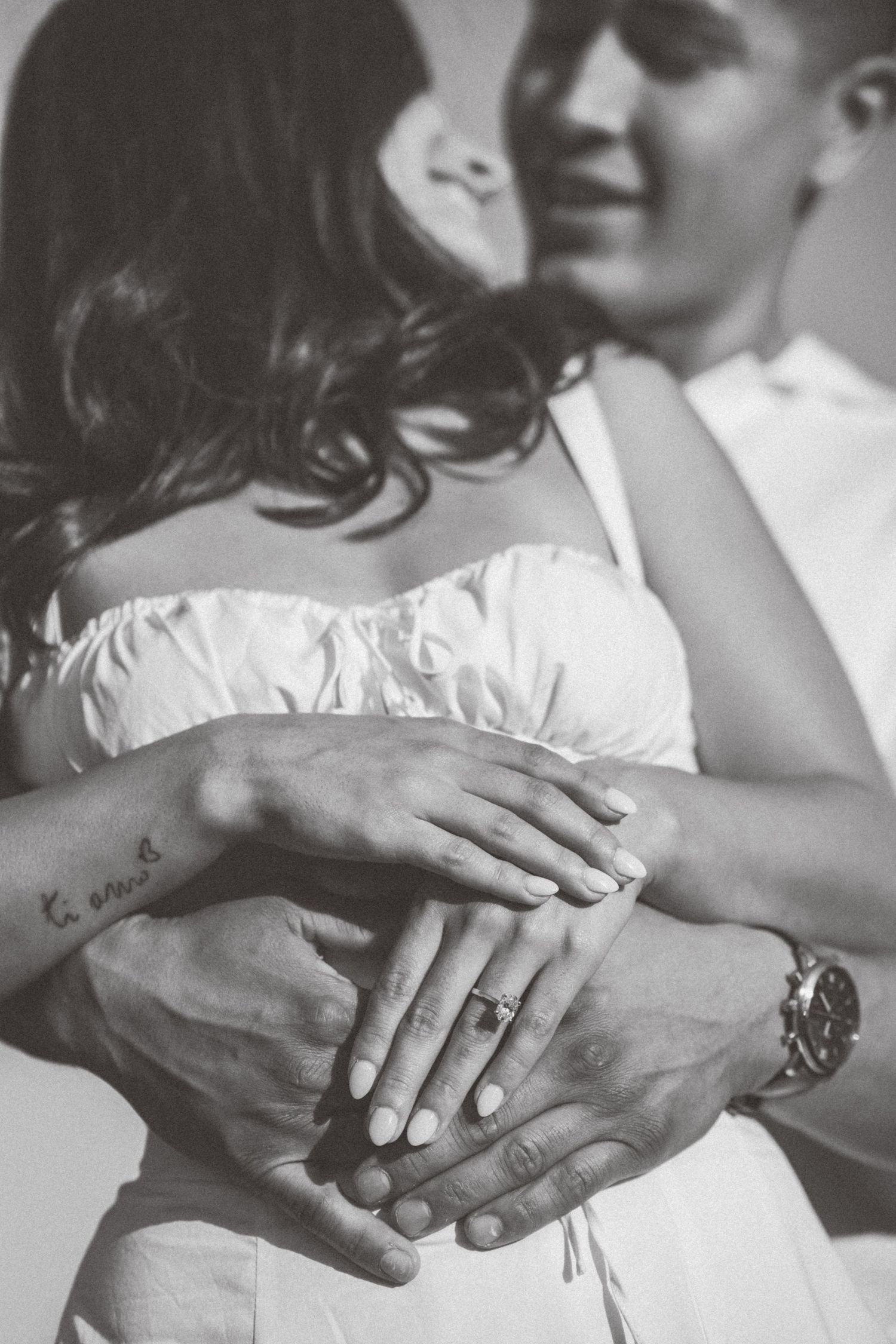 Black and white photo of a couple embracing, showing their hands with an engagement ring, tattoos, and a watch.