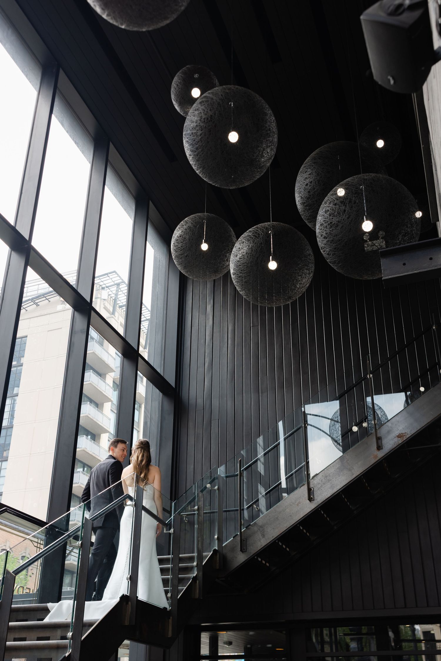 Bride and groom share a romantic moment on modern staircase beneath stylish spherical pendant lights in elegant venue.