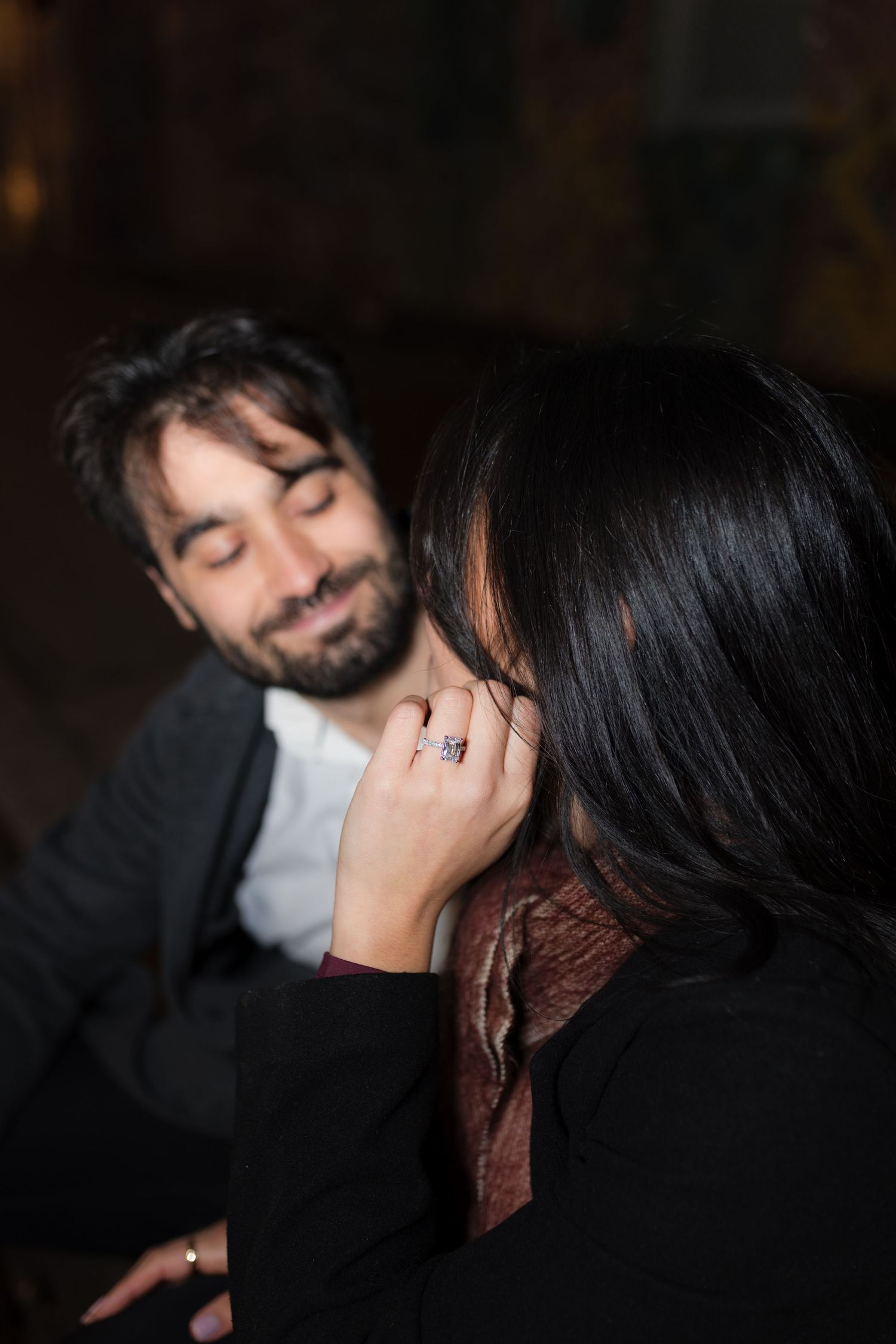 Woman admiring engagement ring while smiling man looks on in romantic dimly lit setting.