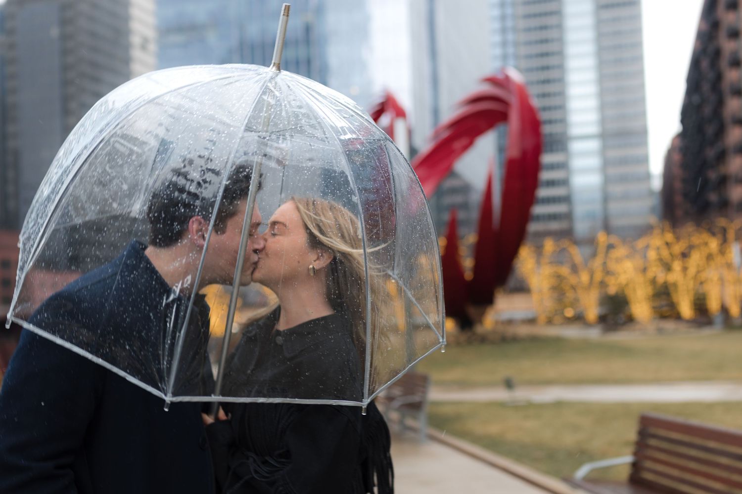 Couple kissing under a clear bubble umbrella in the rain with a red sculpture in the background.