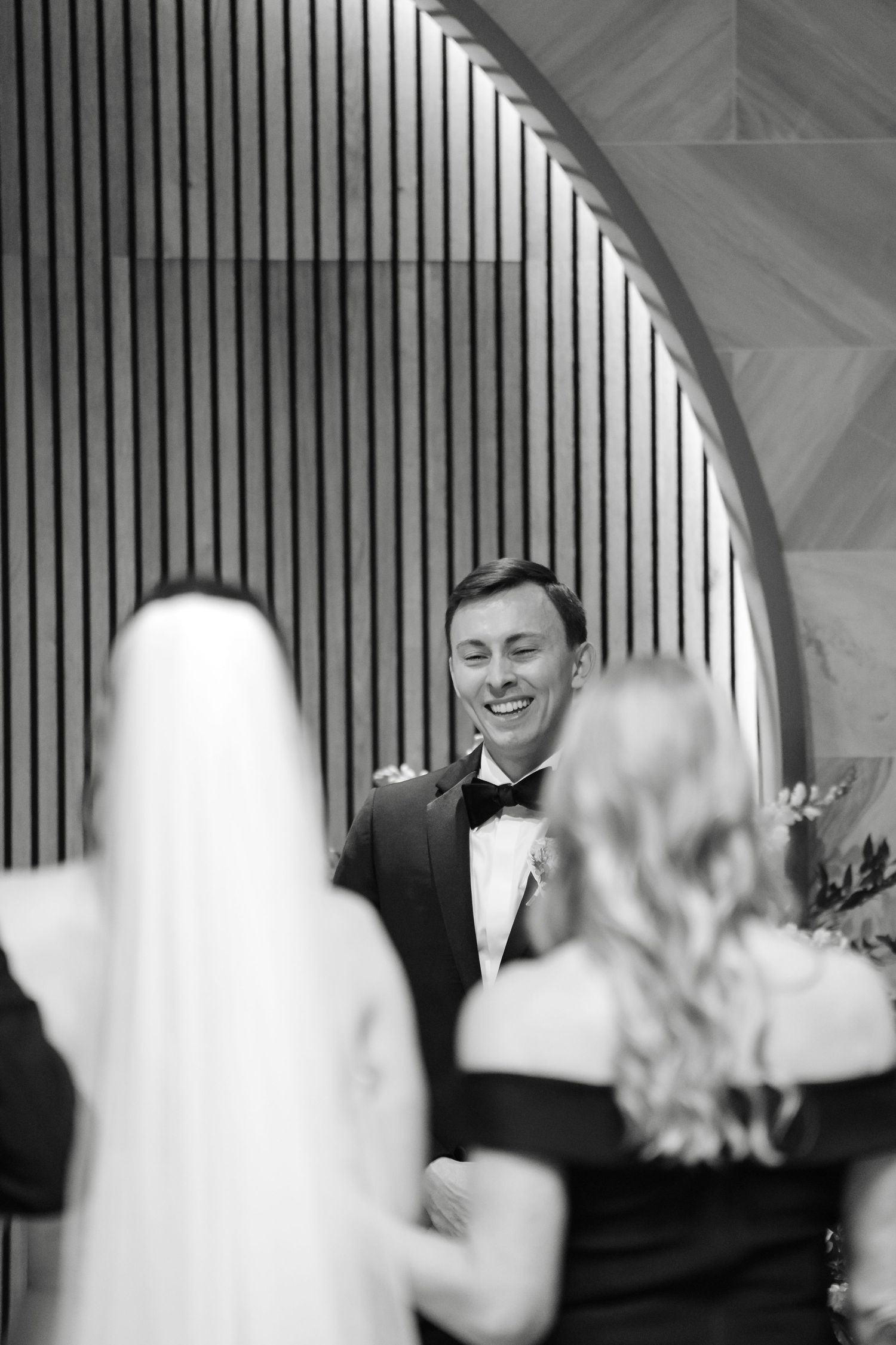 Black and white photo of a groom smiling joyfully as his bride walks toward him at the wedding altar.