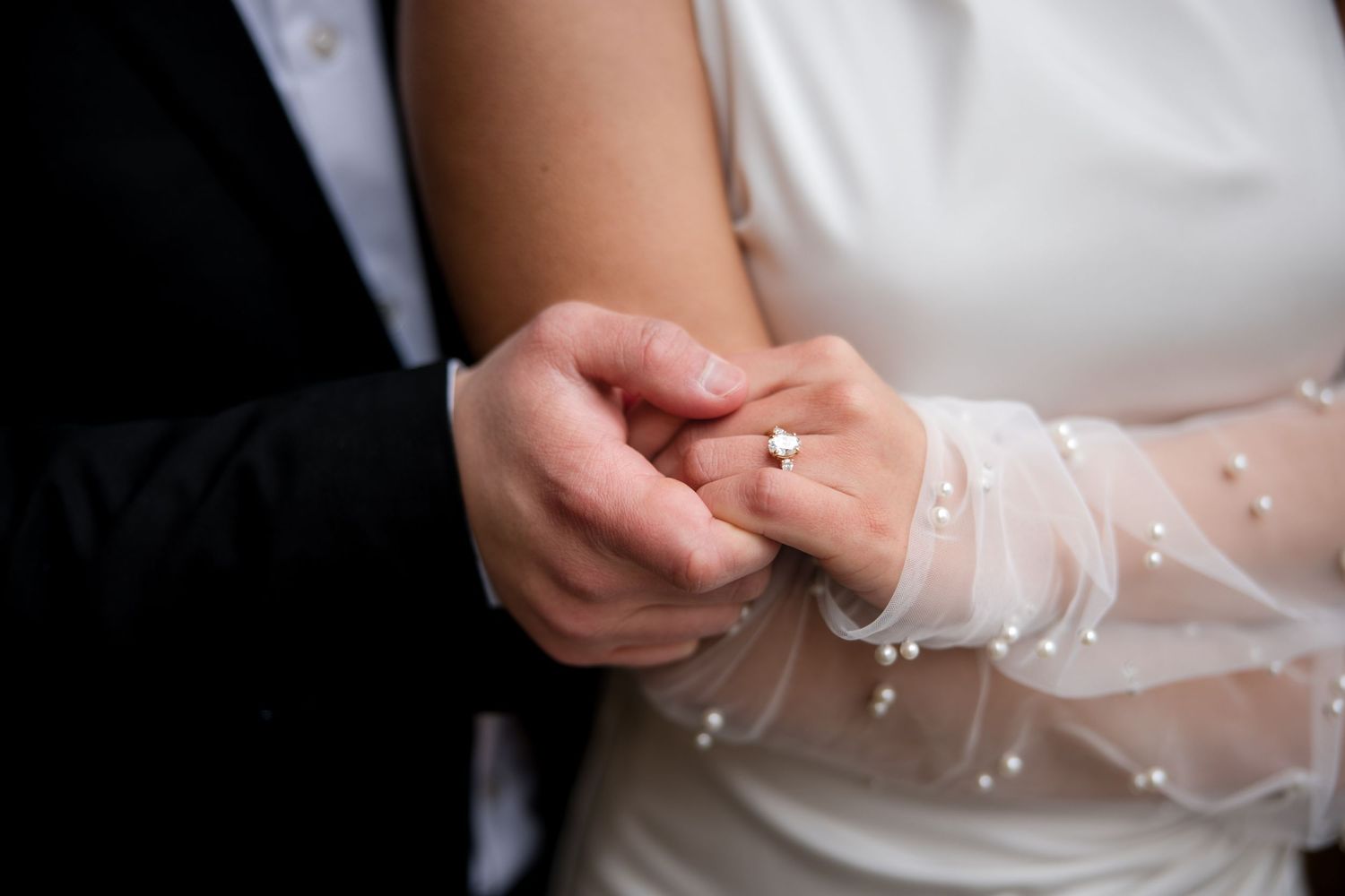 Groom placing a diamond ring on bride's finger during wedding ceremony, bride wearing pearl-adorned white dress.