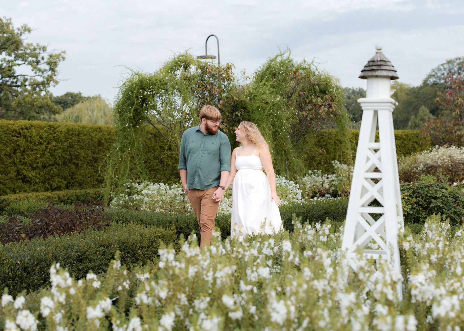 Couple holding hands in a lush garden with white flowers and decorative lighthouse, romantic outdoor engagement photo.