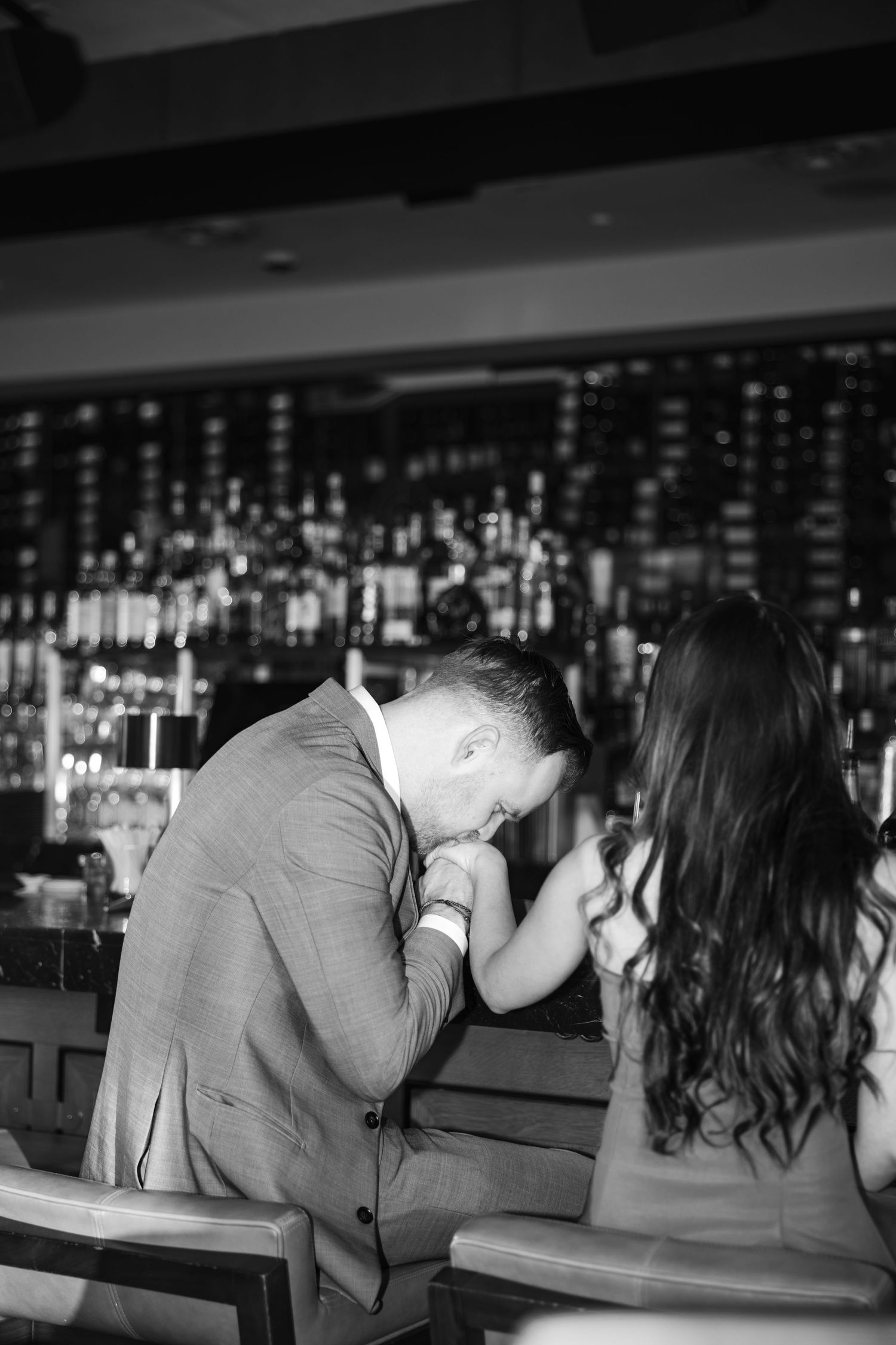 A man in a suit gallantly kisses a woman's hand at an elegant bar in this romantic black and white photo.