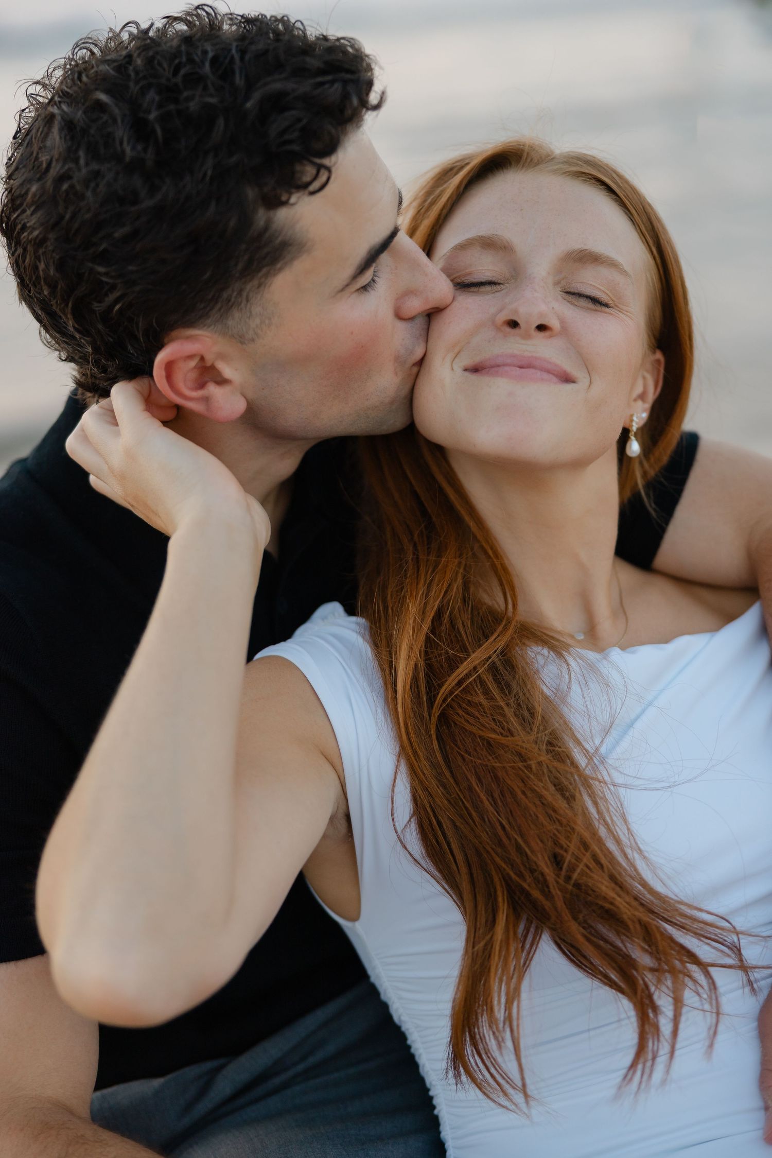 A man with curly hair kisses a smiling redhead woman on the cheek outdoors.