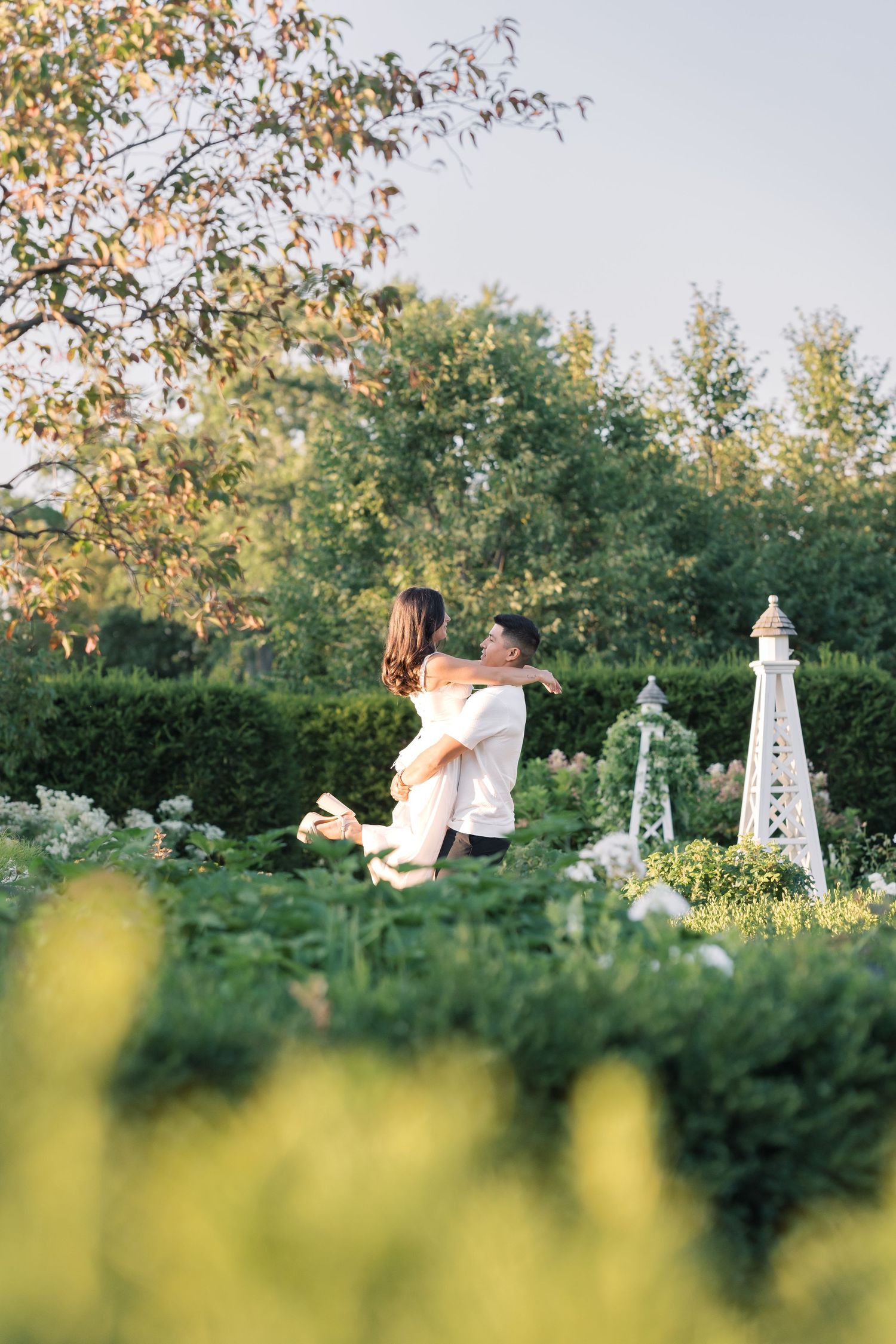 Couple embracing in a lush green garden with hedges and a decorative white tower during golden hour.