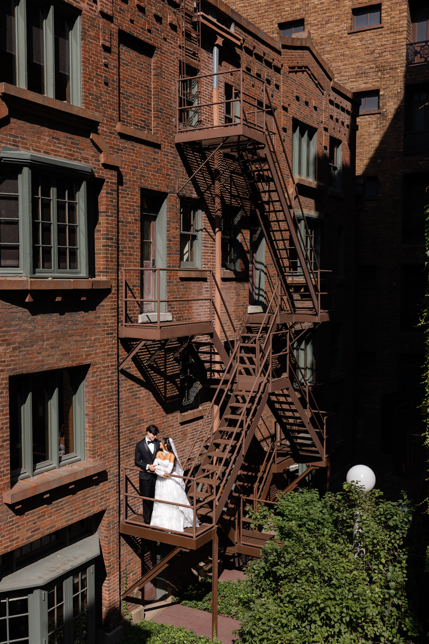 Bride and groom pose romantically on a rustic iron fire escape of a brick building surrounded by lush greenery.