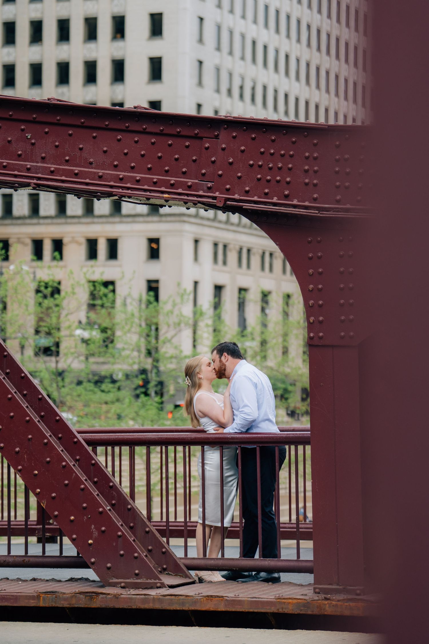 Couple kissing on a red iron bridge in Chicago with city buildings in the background.