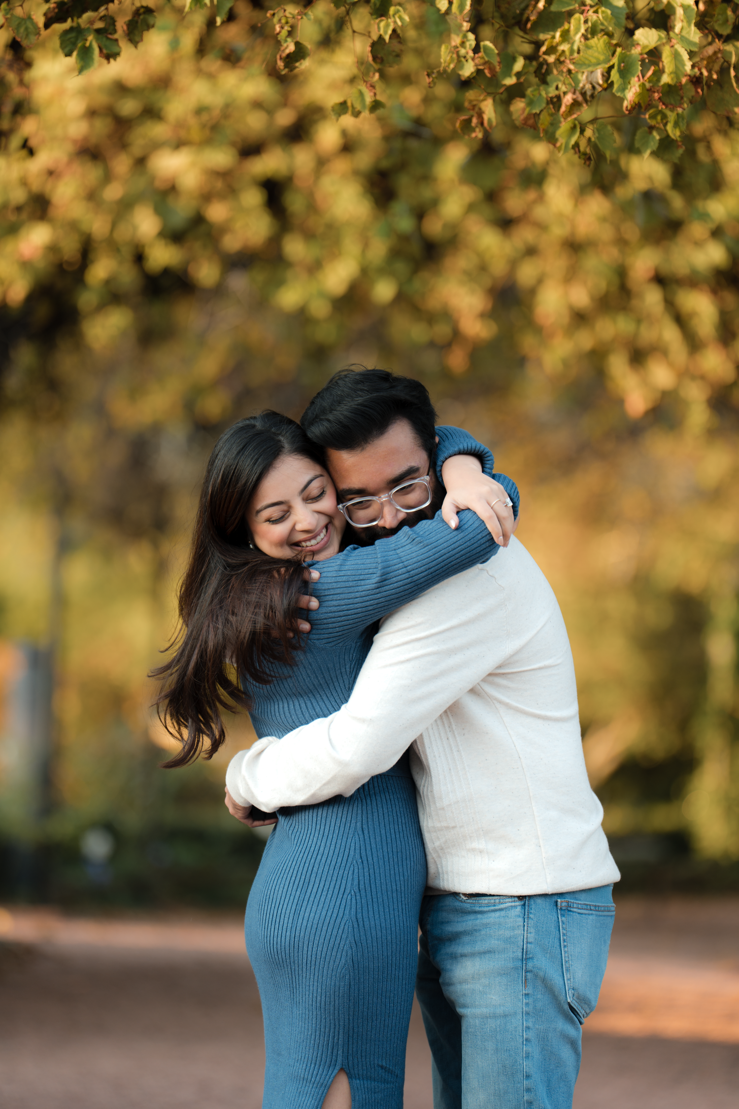 A couple shares a warm embrace outdoors in autumn, surrounded by golden yellow foliage in a park setting.