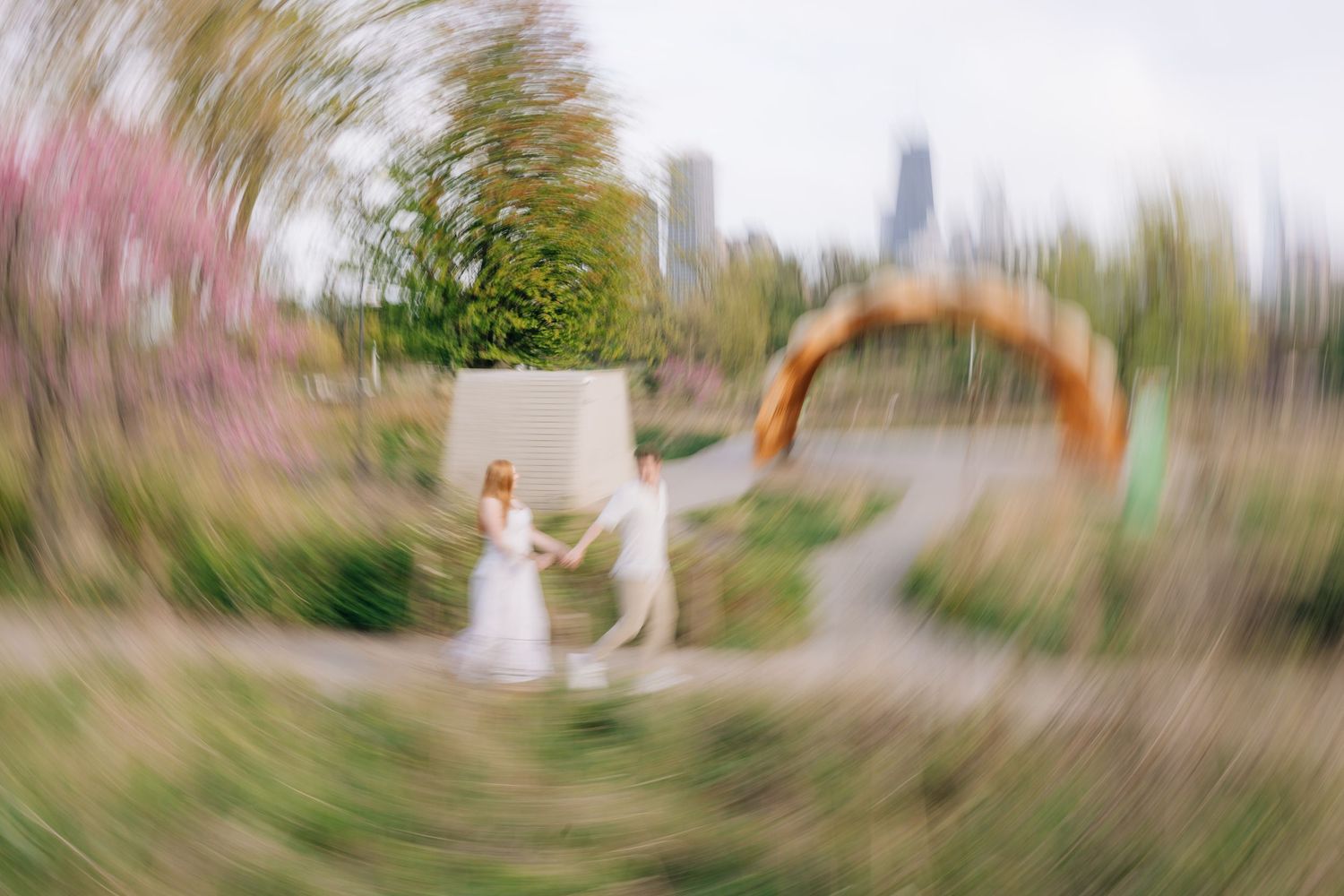 Couple in wedding attire dancing in a lush garden with a city skyline, captured with a dreamy zoom blur effect.