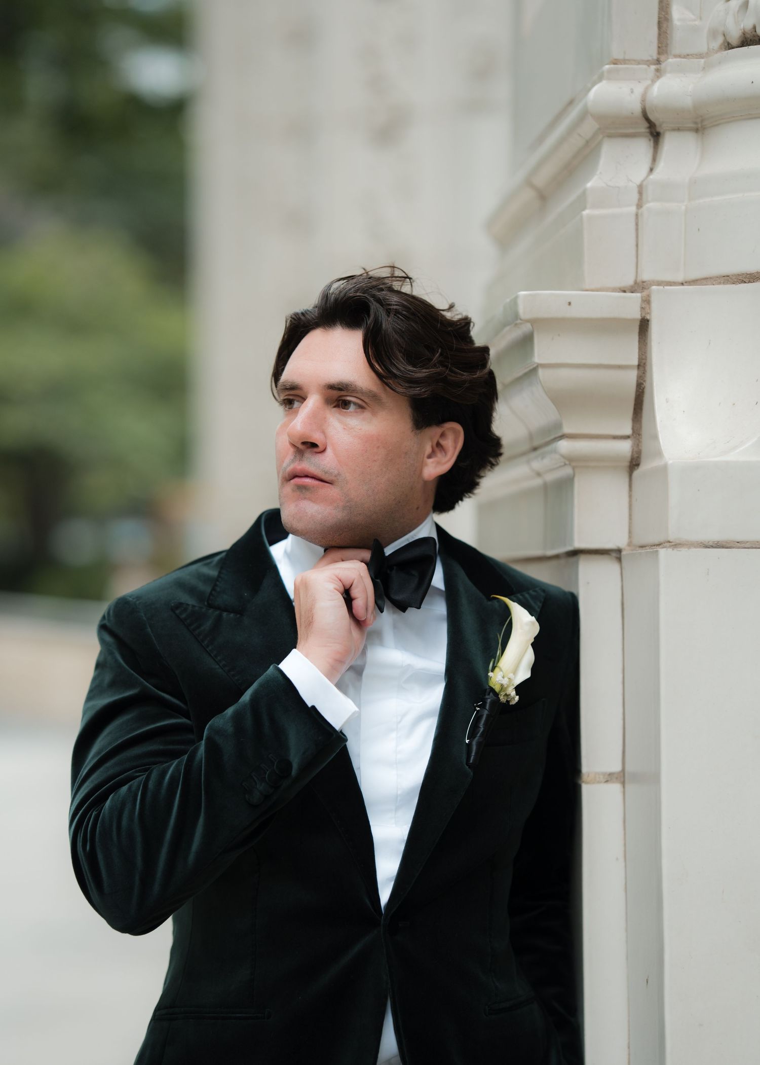 Groom in black tuxedo poses thoughtfully against white stone column.