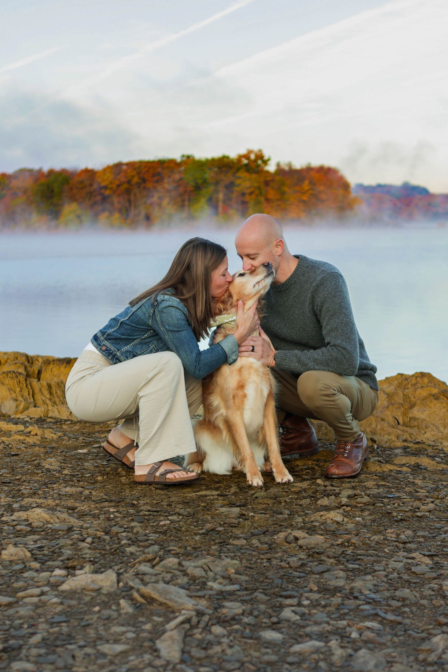 Couple kissing their golden retriever dog on rocky shore with misty autumn lake and colorful trees behind.