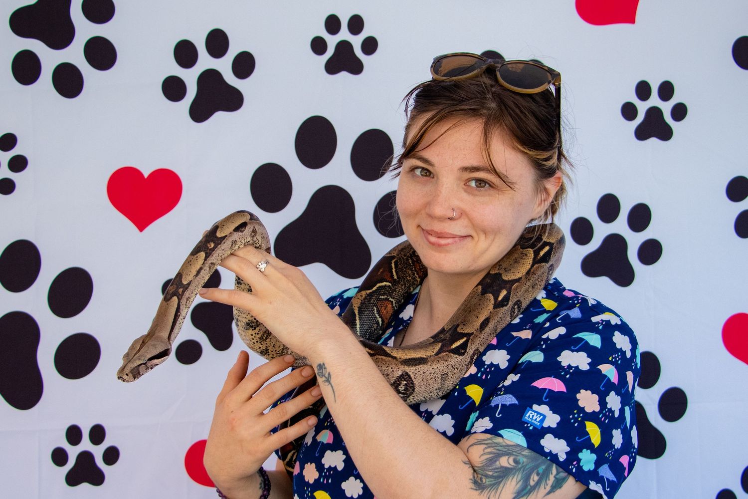 Smiling young woman with sunglasses holding a boa constrictor snake in front of a paw print backdrop.