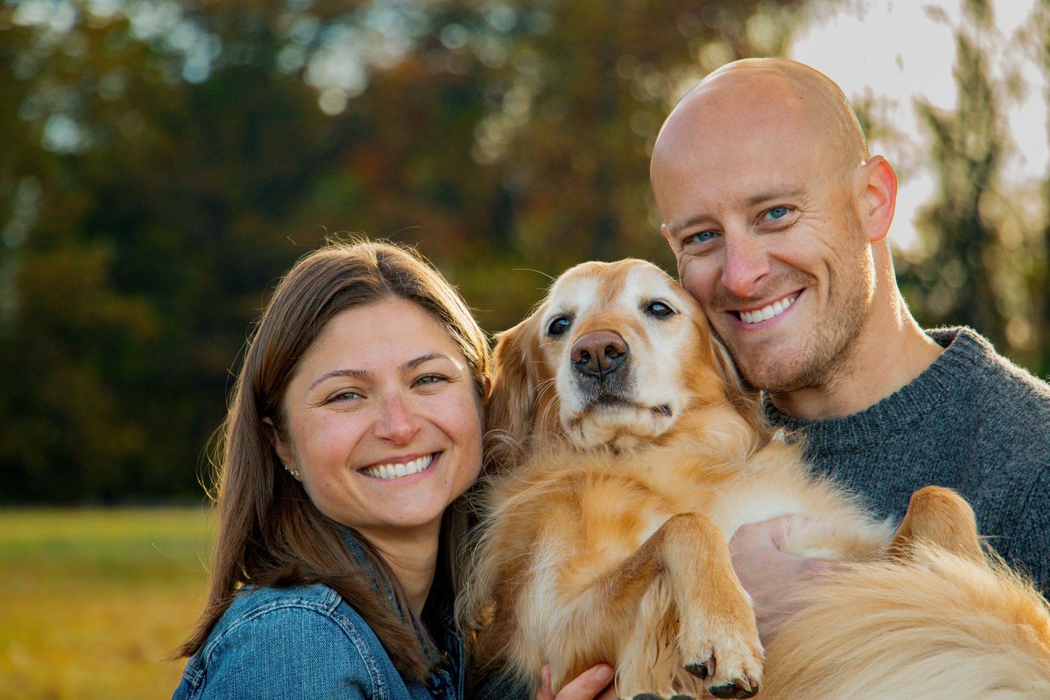 Smiling couple embracing their happy golden retriever dog outdoors in a warm autumn park setting.