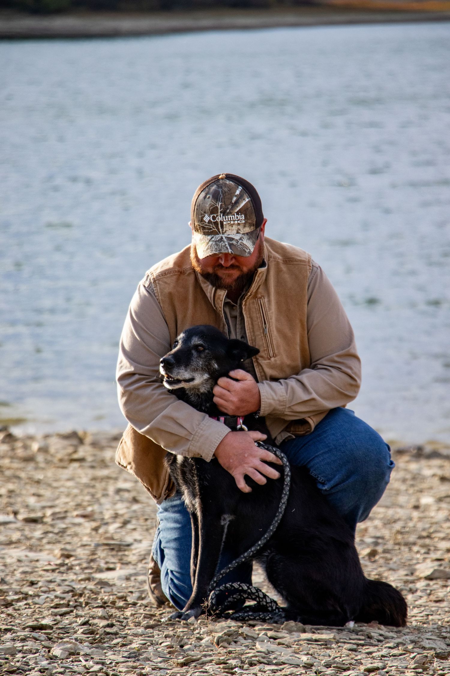 Man in camo cap kneeling on rocky shore tenderly embracing his senior black dog near a lake.