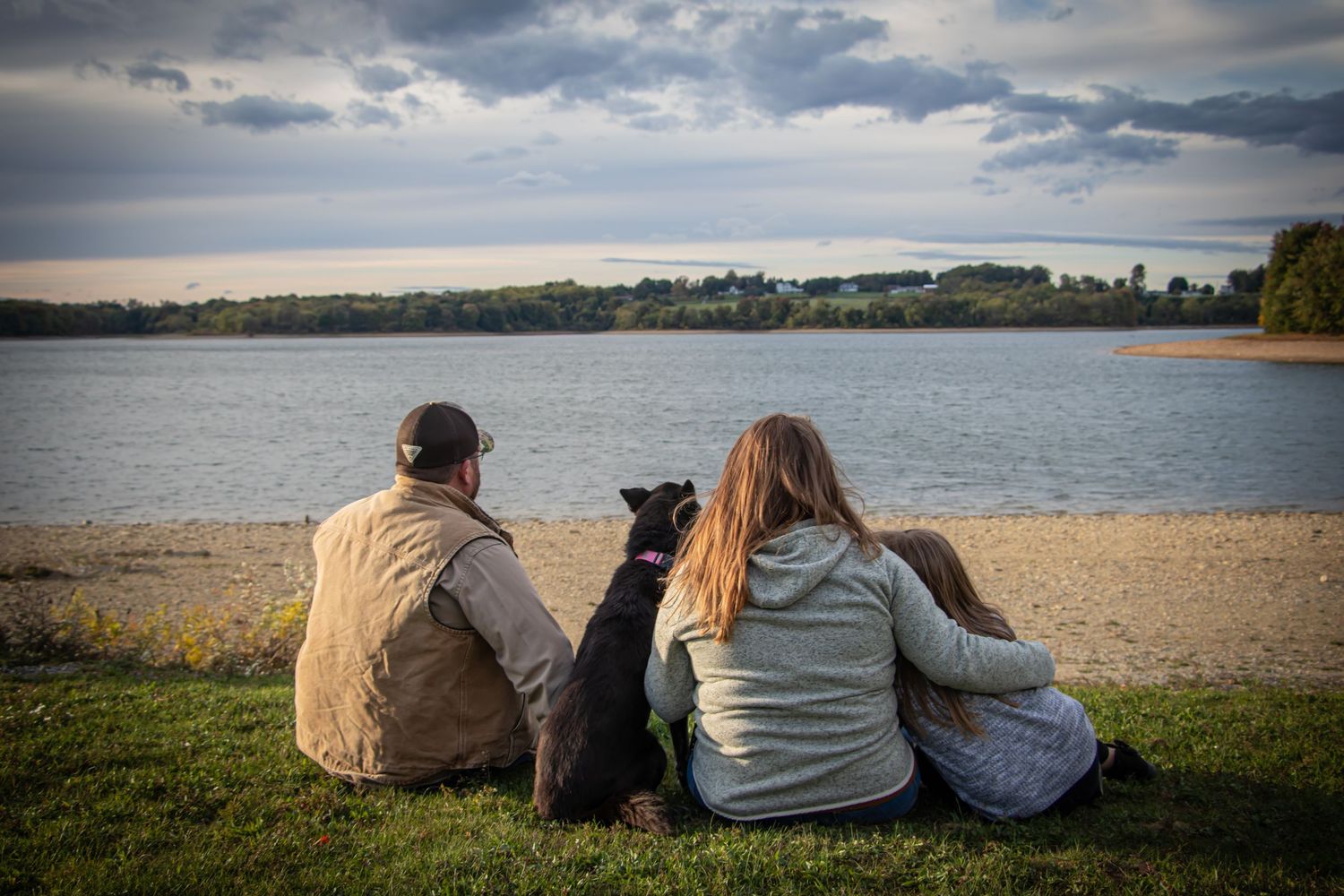 Couple sitting with their black dog on a grassy lakeside shore enjoying a peaceful cloudy sunset view.