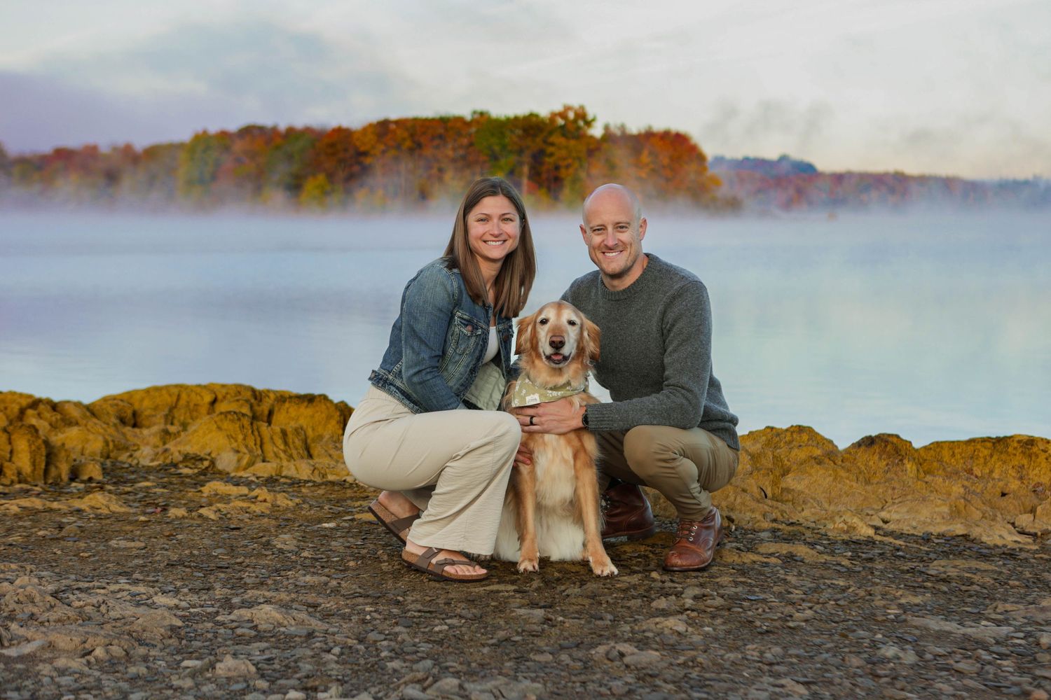 Couple crouching with their golden retriever dog by a misty lake surrounded by colorful fall foliage.