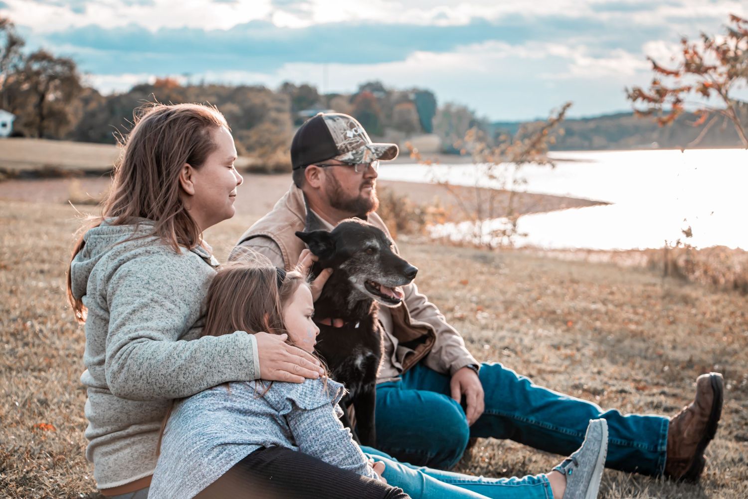 Family with young child and black dog sitting together on grass by a lake on autumn day.