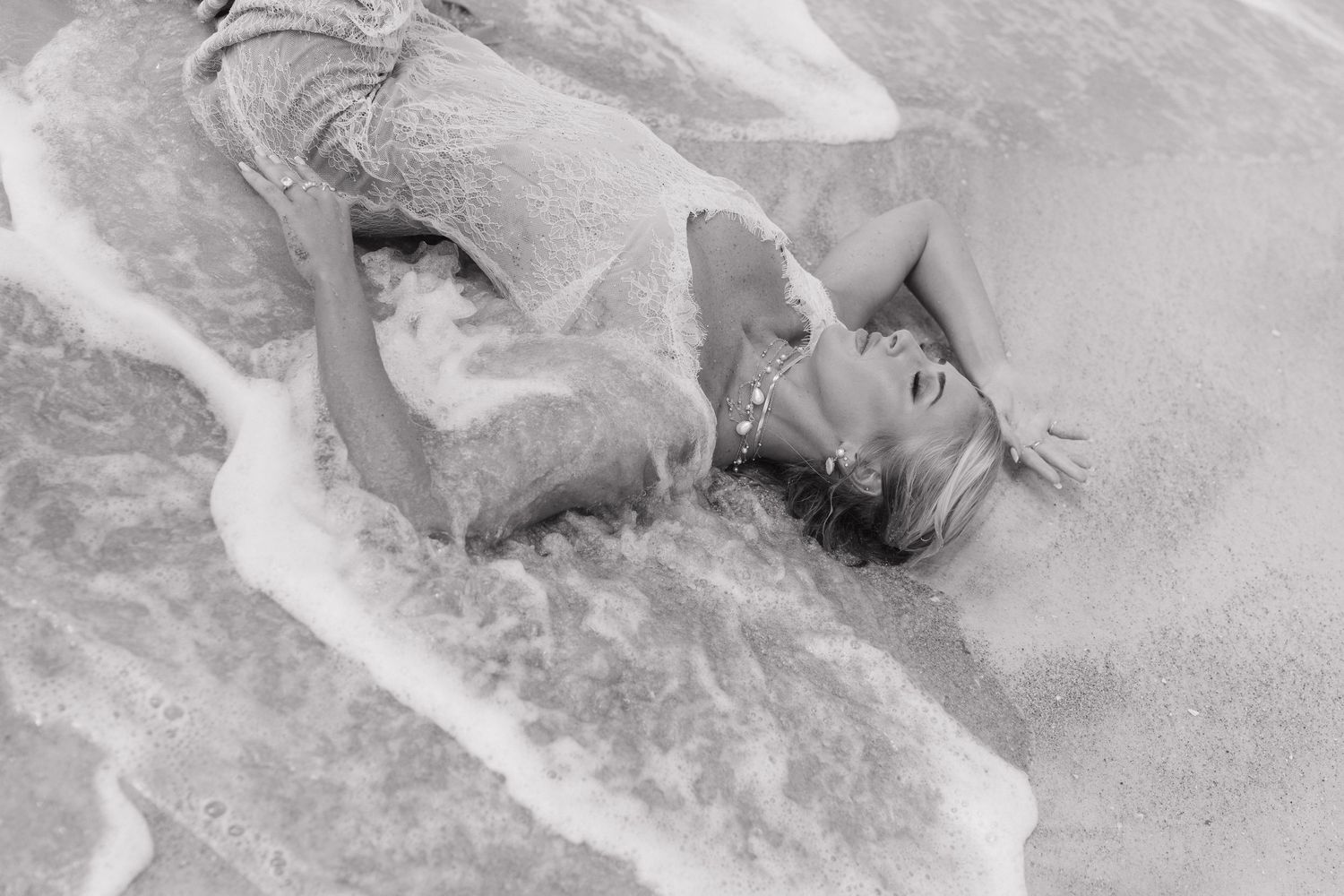 Black and white photo of a woman lying in ocean waves on a sandy beach, wearing a sparkly outfit.