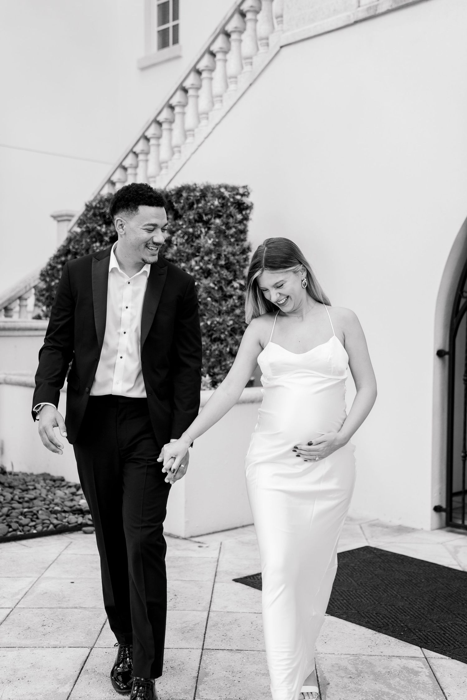 Black and white photo of newlyweds walking together outdoors, with bride in sleek white gown and groom in dark suit.