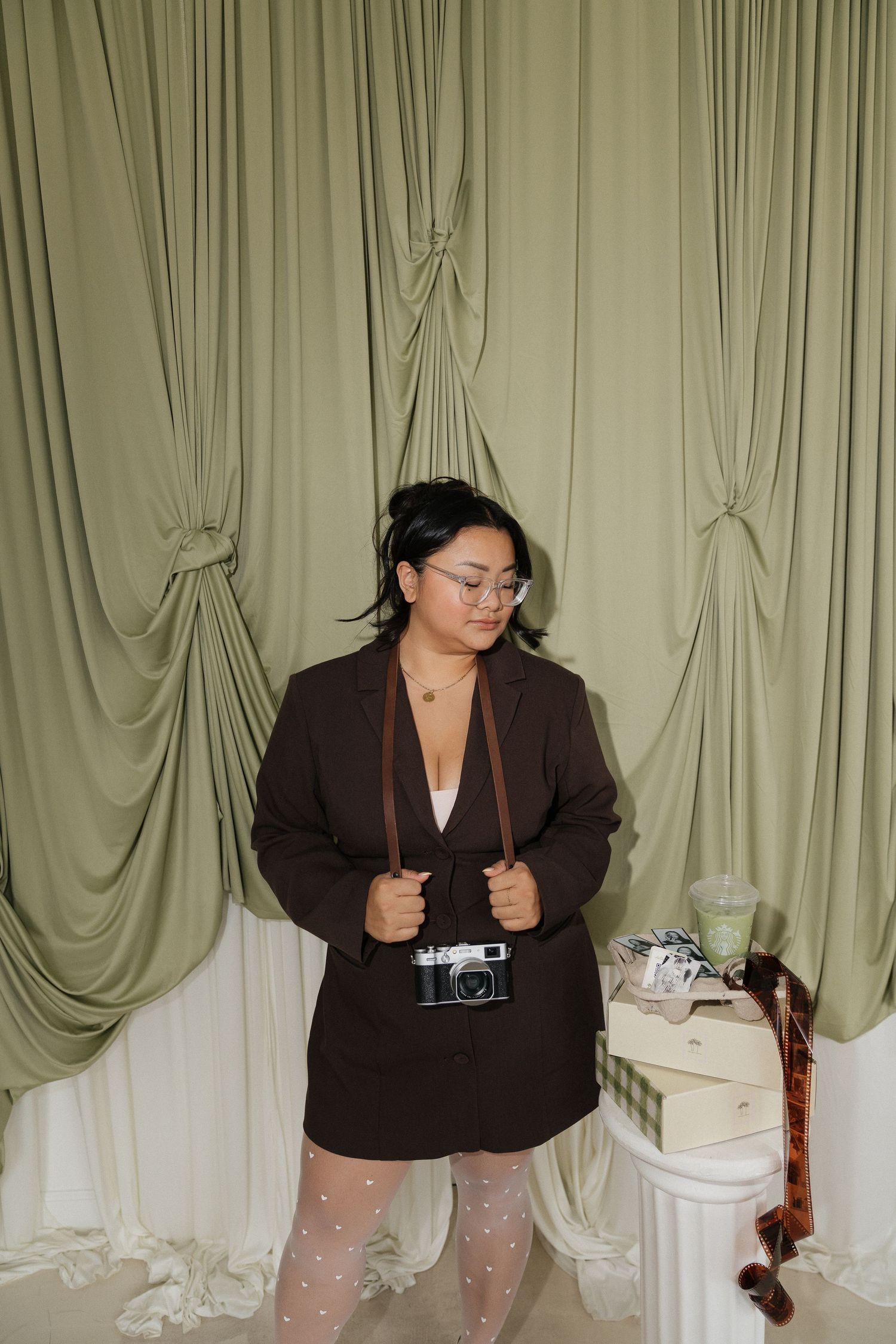 Woman in brown blazer and skirt with film camera poses before sage green draped curtains.