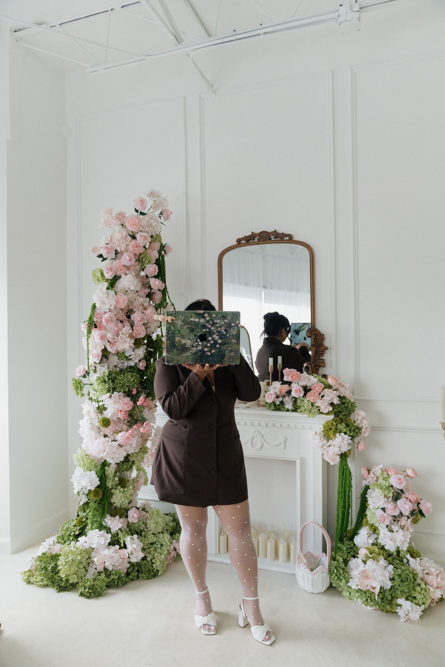 Person holding artwork in front of face near floral arrangements and ornate mirror in white room.