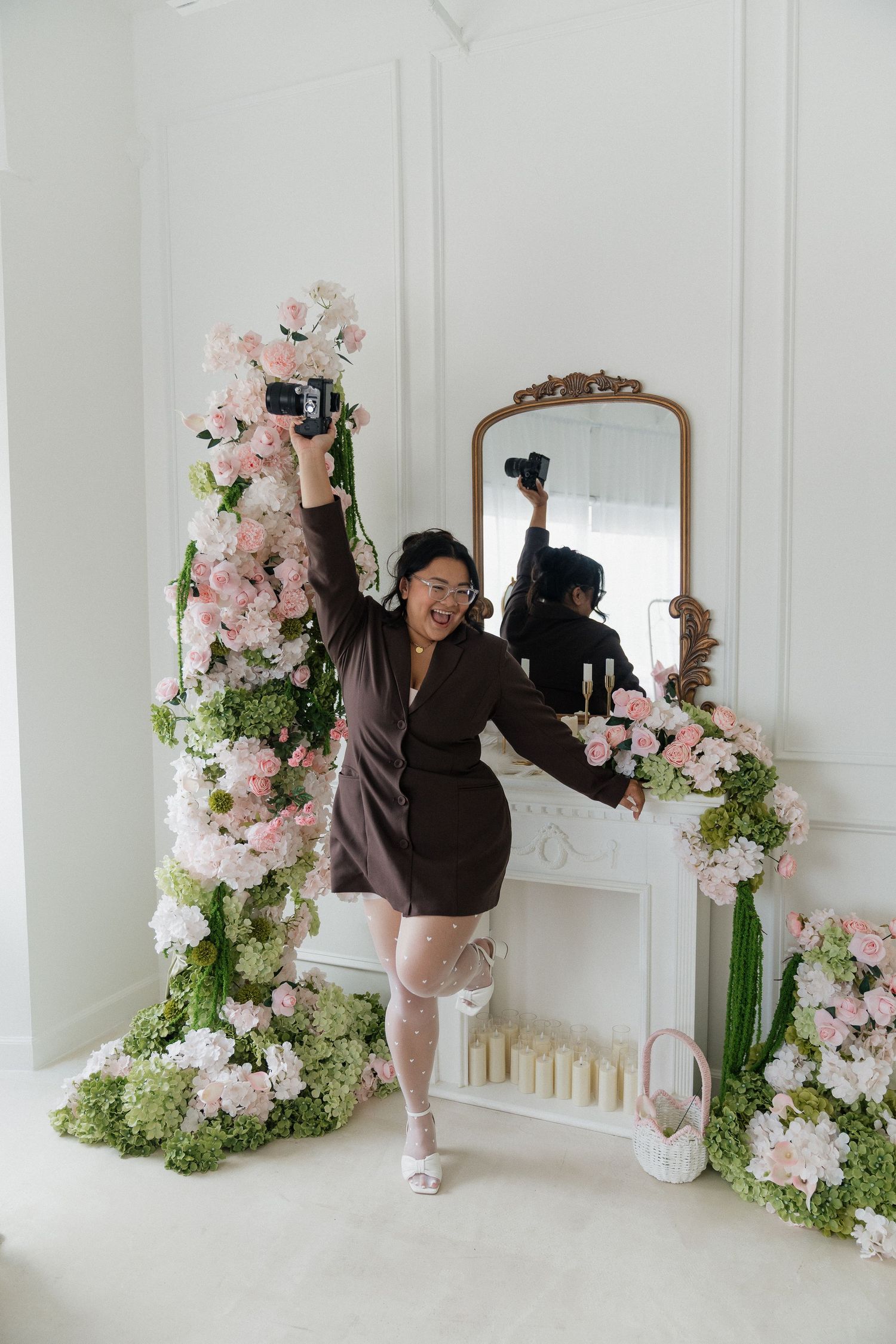 Woman in brown dress takes selfie with camera among pink floral arrangements beside ornate mirror and fireplace.
