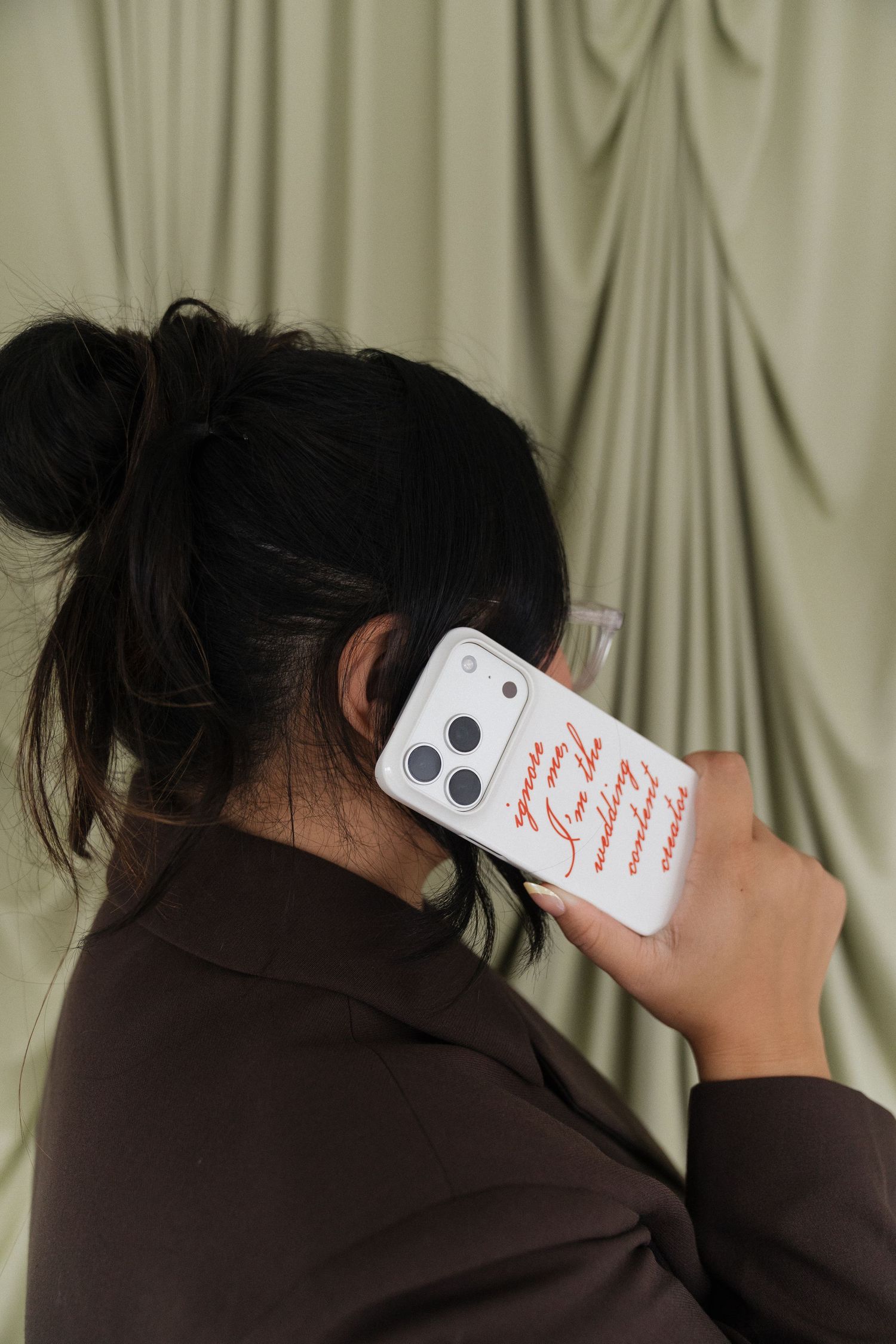 Woman holding white iPhone case with red script text up to her ear, wearing brown blazer against sage green draped backdrop.