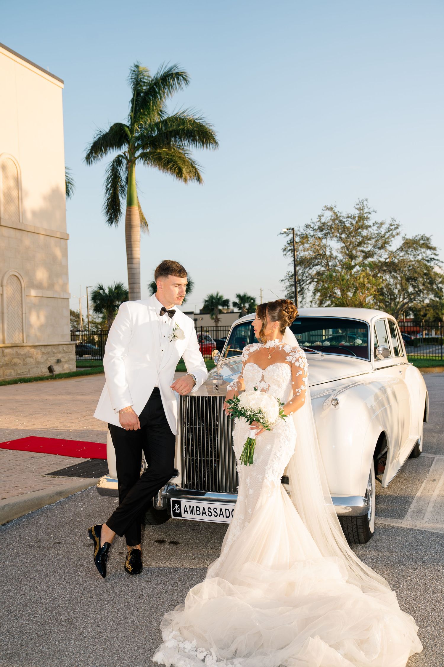 Elegant wedding couple poses next to vintage white Rolls Royce with palm tree and red carpet in background.