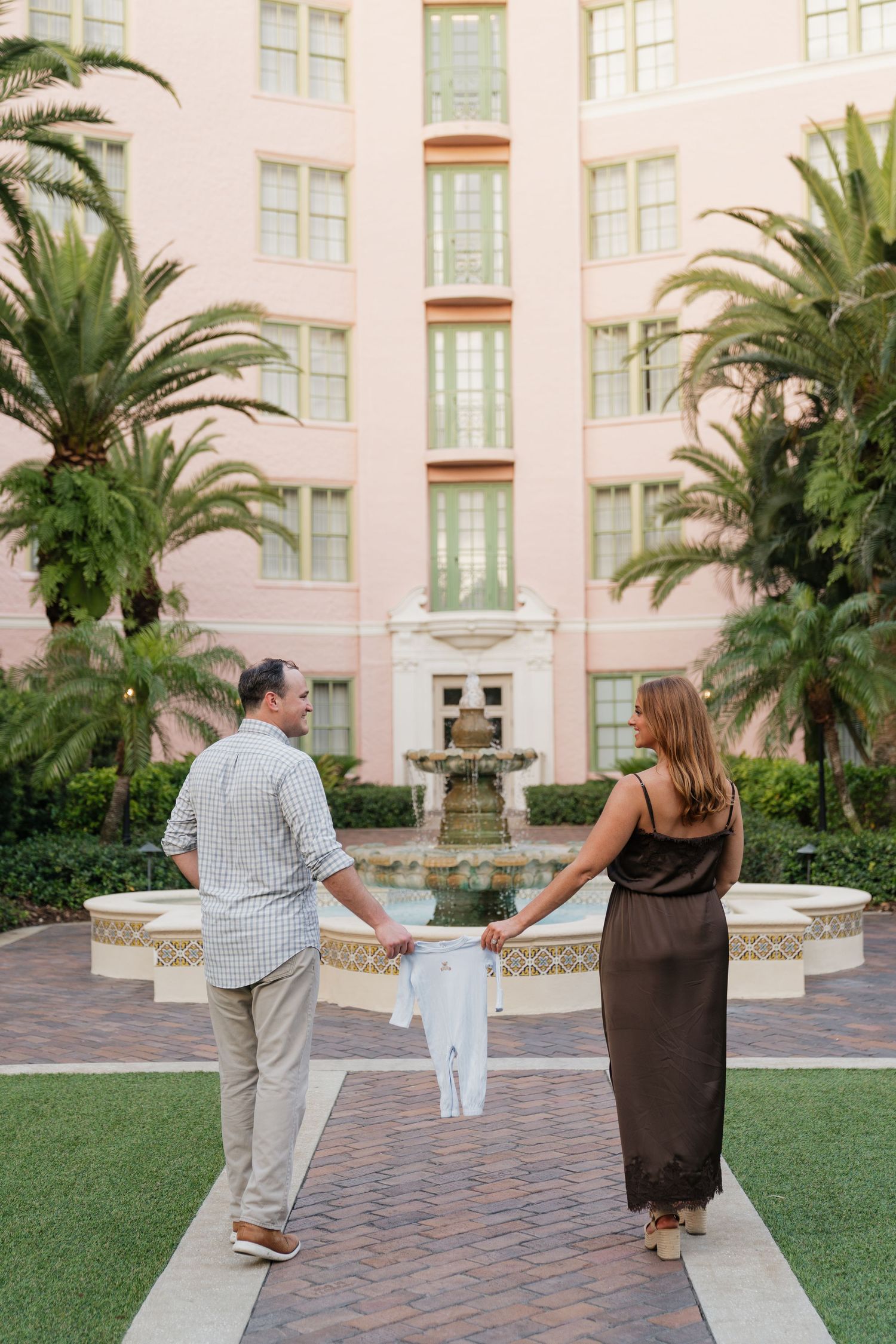 Couple holding baby onesie between them in front of a pink hotel fountain surrounded by palm trees.