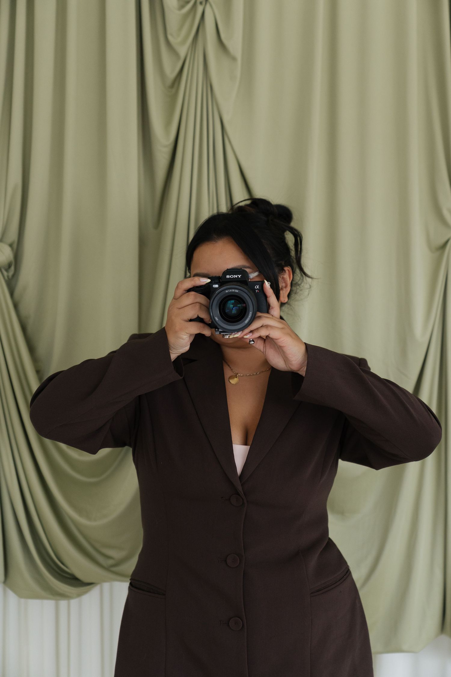 Woman in brown blazer photographing with DSLR camera in front of sage green draped curtain backdrop.