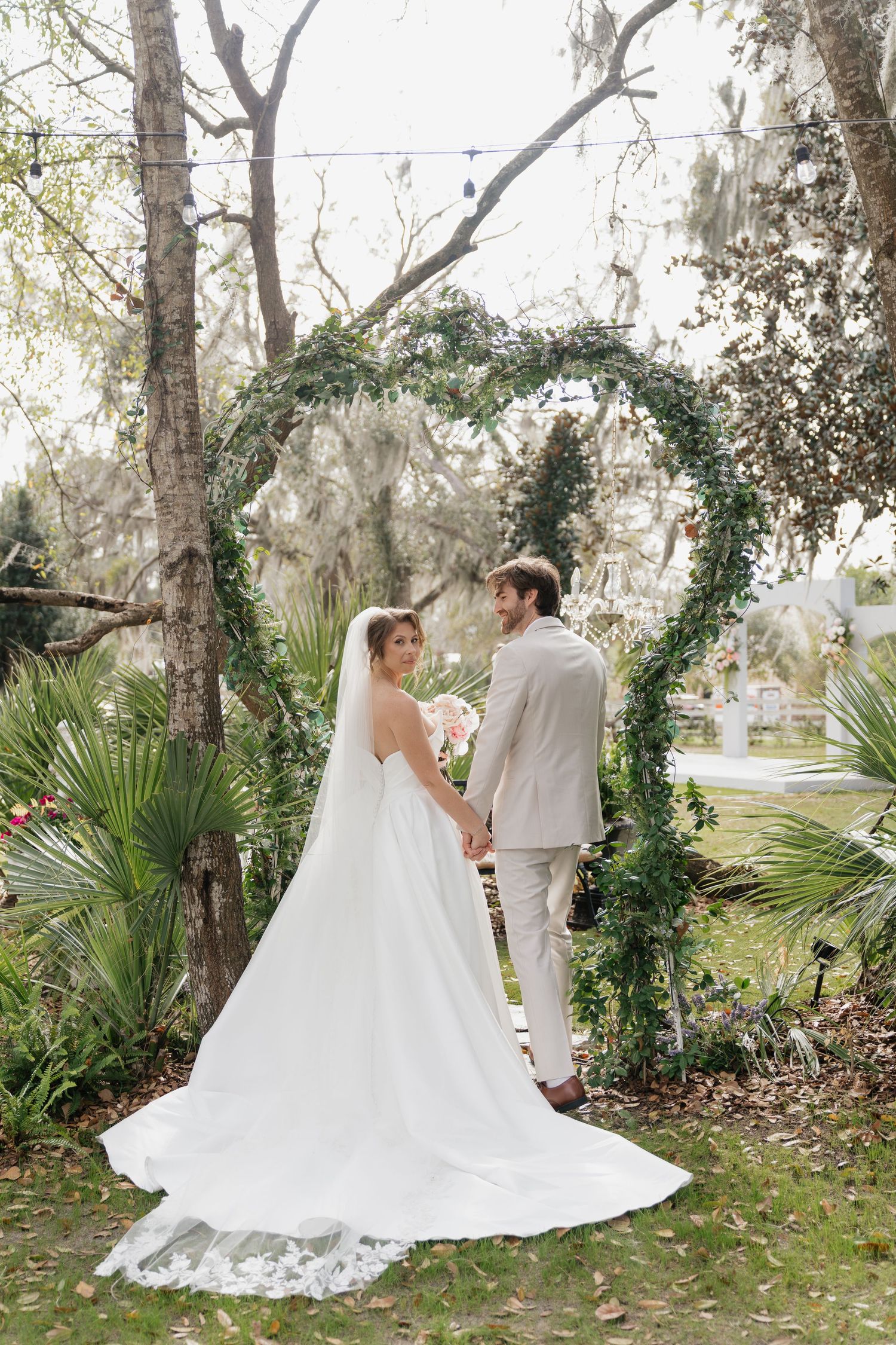 A bride and groom stand under a lush green floral arch in a garden setting for their wedding ceremony.