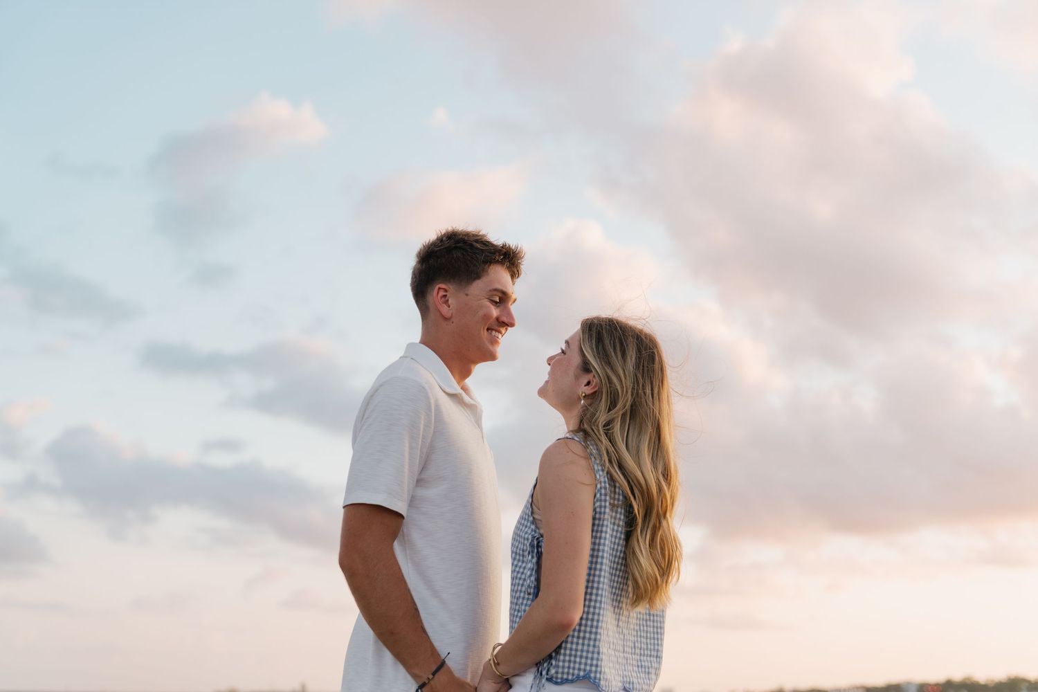 Couple holding hands and smiling at each other outdoors at sunset with pastel pink cloudy sky background.