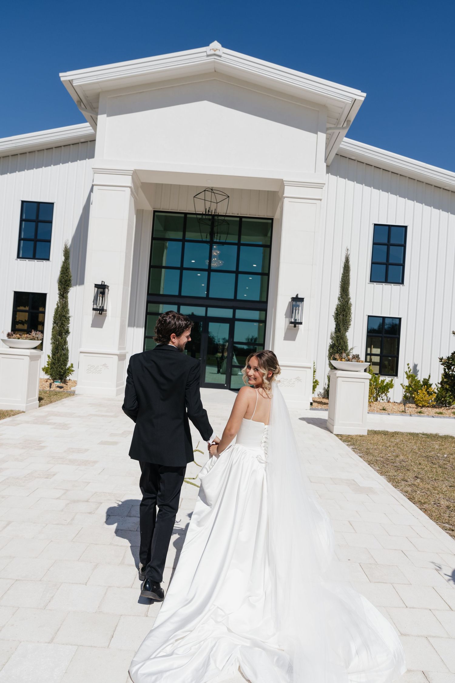 A couple in formal wedding attire walks away from the camera towards a modern white church building on a sunny day.