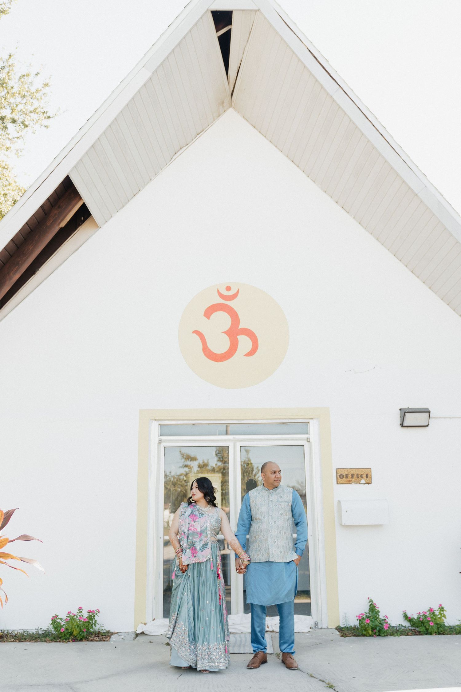 A couple stands at the entrance of a white building with an orange Om symbol above the door.
