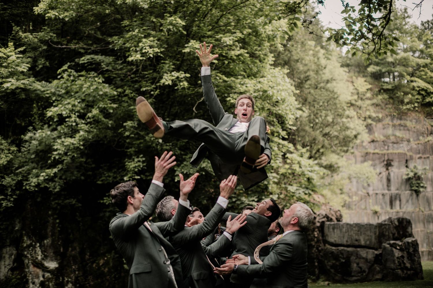 Groomsmen toss groom in air during playful outdoor wedding photo.