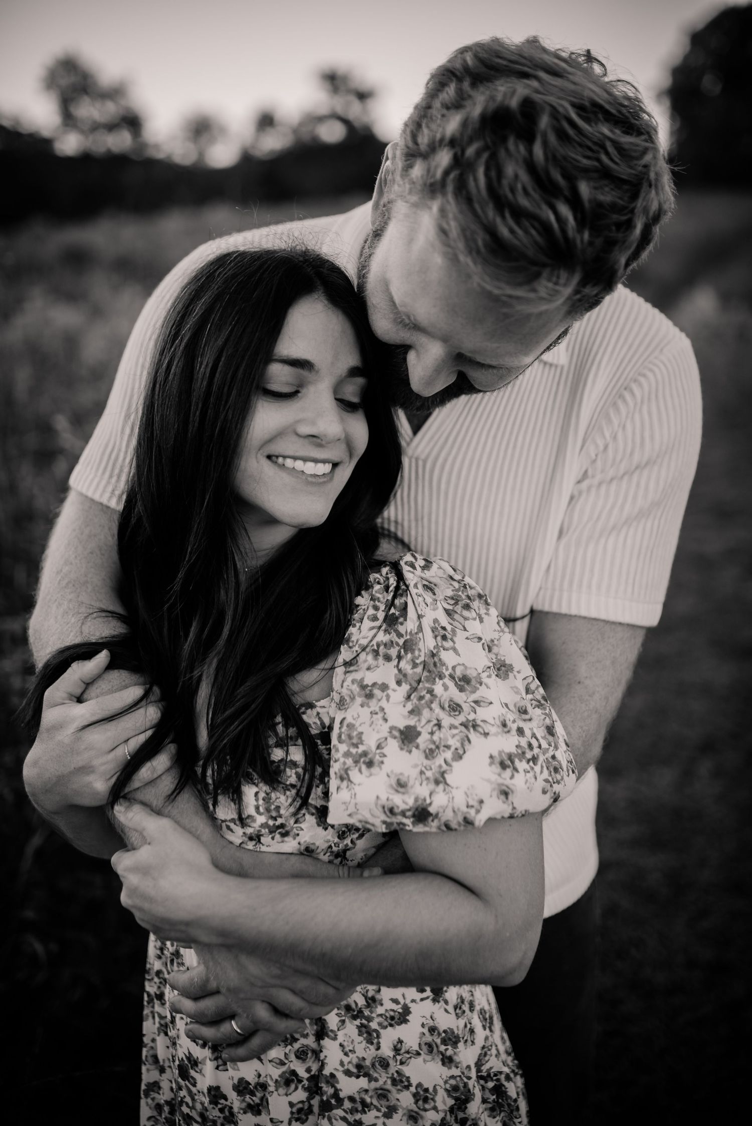 A romantic black and white photo series of a couple embracing outdoors in a field at sunset.