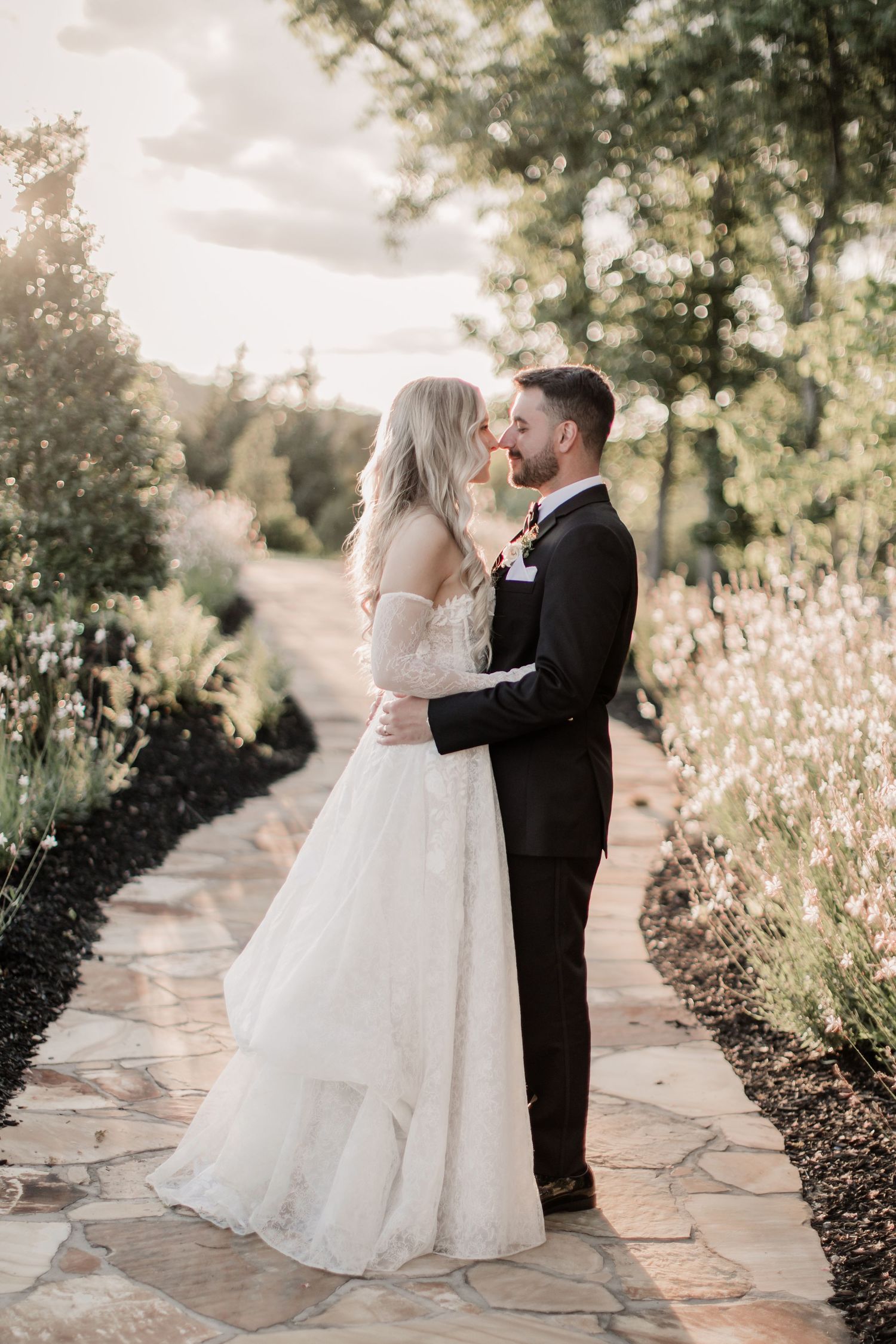 A couple shares an intimate moment on a stone path surrounded by lush garden foliage in romantic wedding attire.