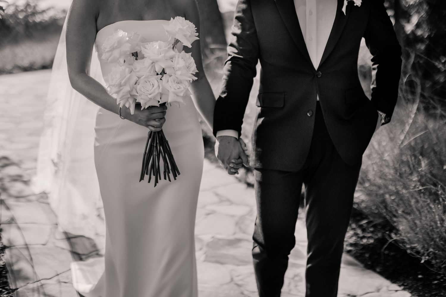A black and white wedding photo showing a couple walking together outdoors while holding a bouquet of flowers.
