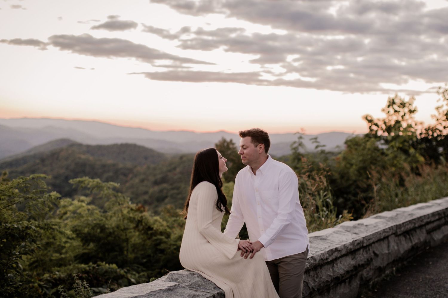 A couple shares a romantic moment at sunset overlooking a scenic mountain landscape from a stone wall viewpoint.