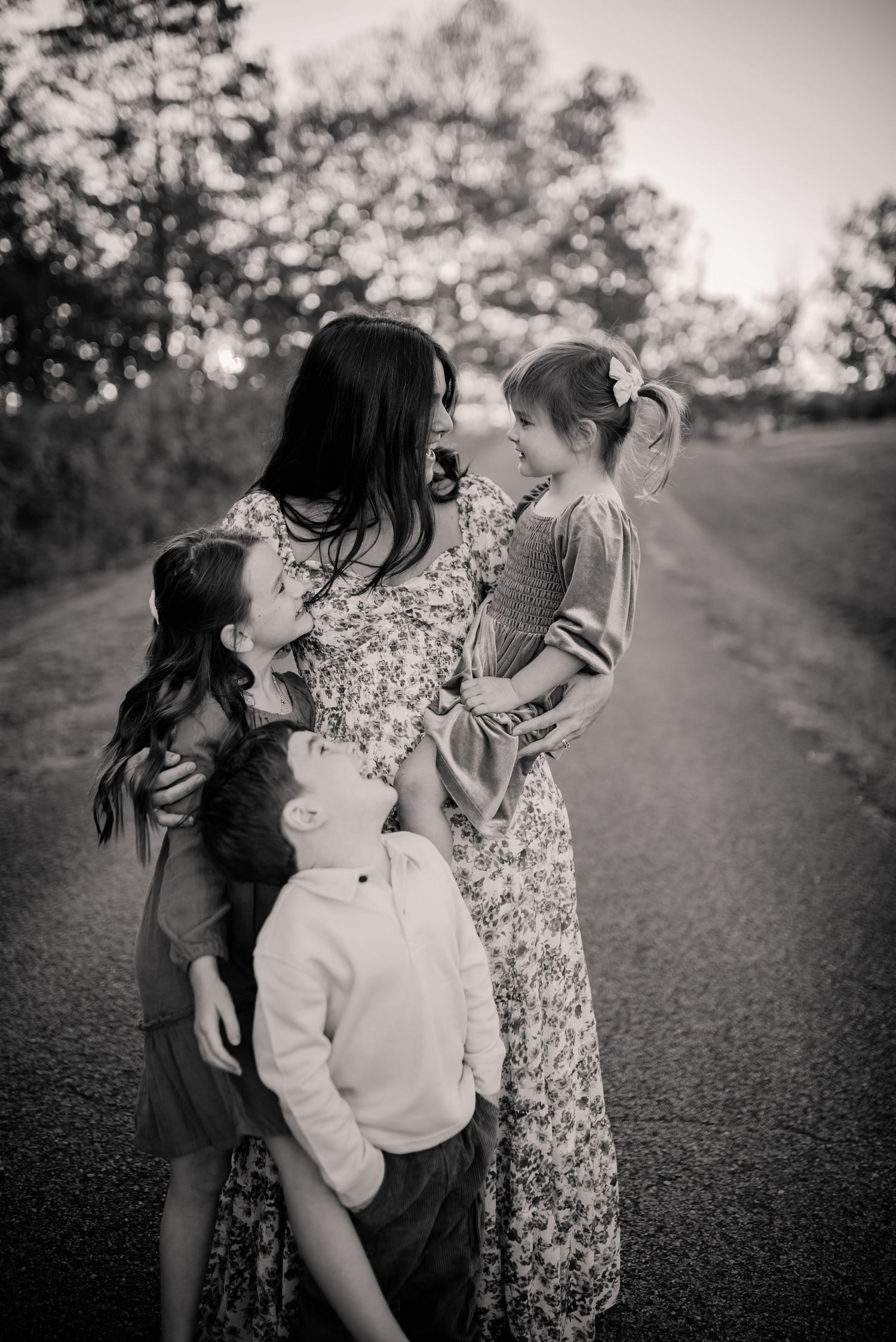 A family shares a tender moment together on a tree-lined path during golden hour in black and white.