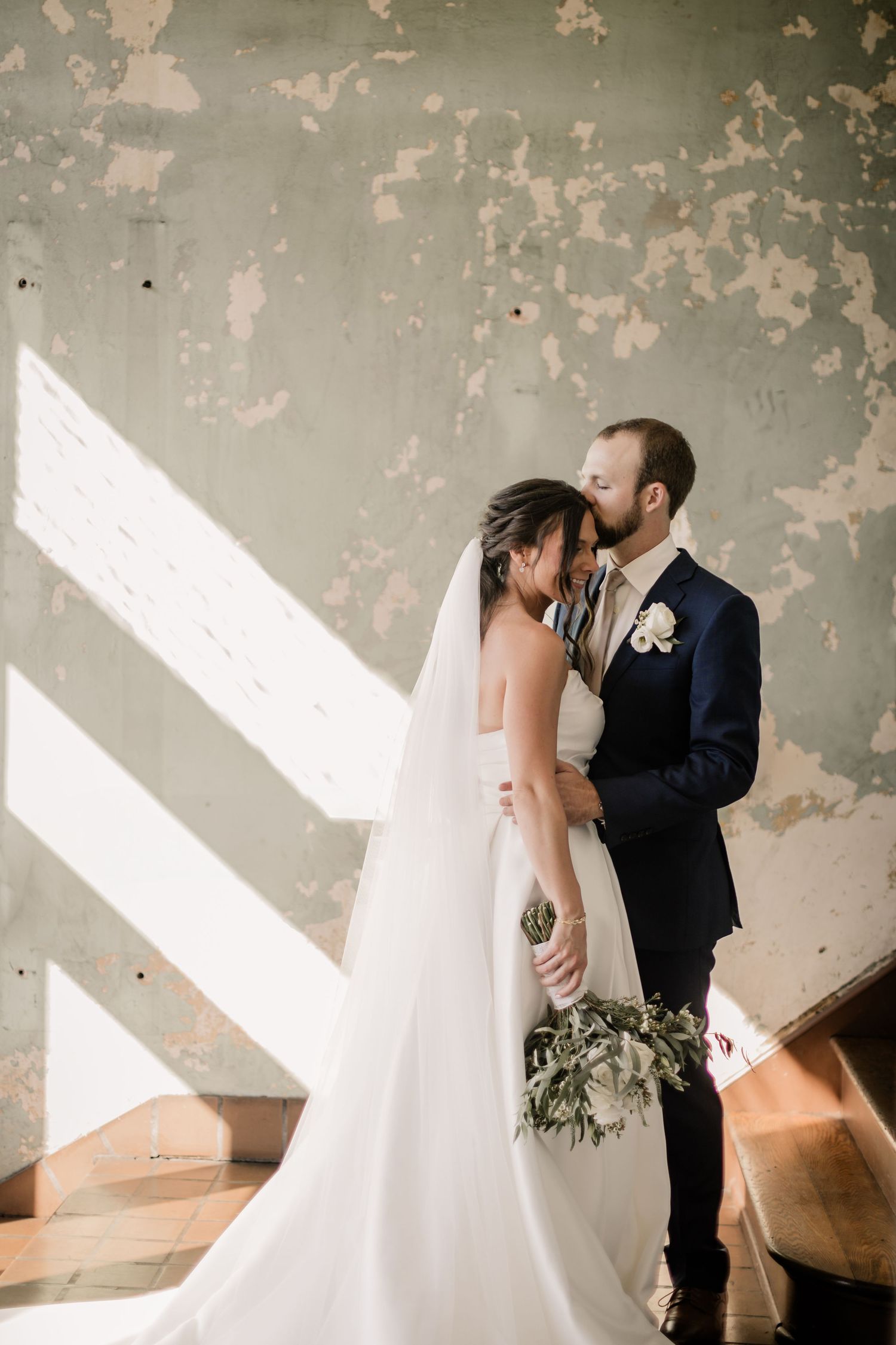 Elegant couple shares tender moment against weathered wall with dramatic lighting.