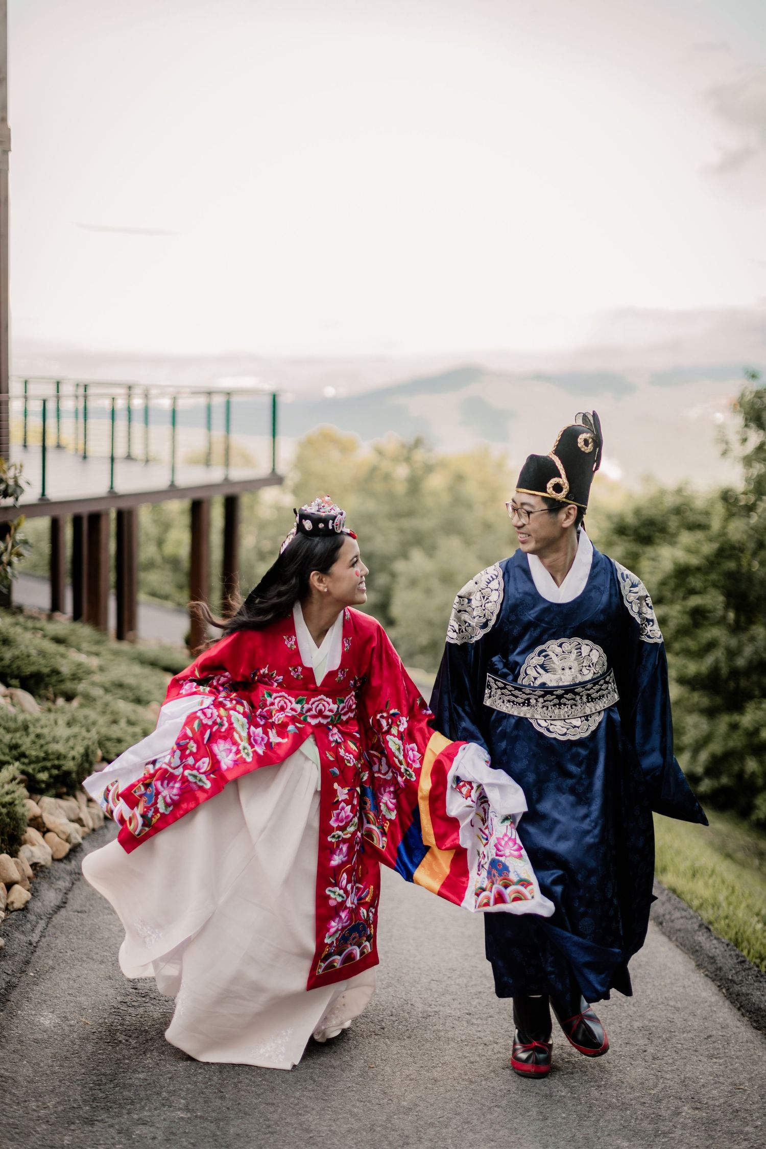 A couple wearing traditional Asian wedding attire walks together on a scenic mountain path.