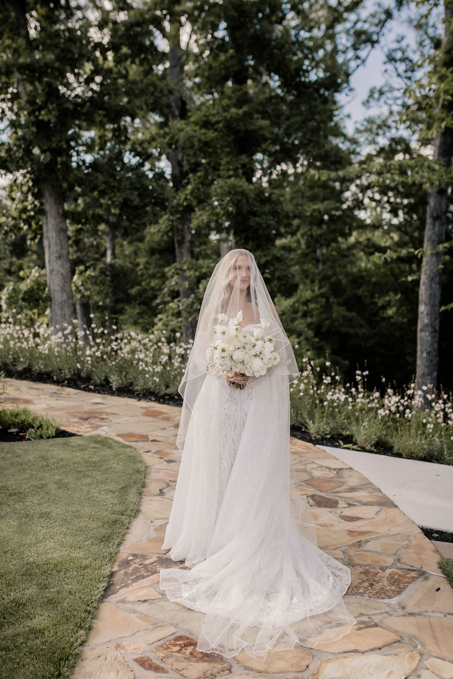 A bride in a flowing white wedding gown with a long veil stands on a stone path surrounded by lush greenery.