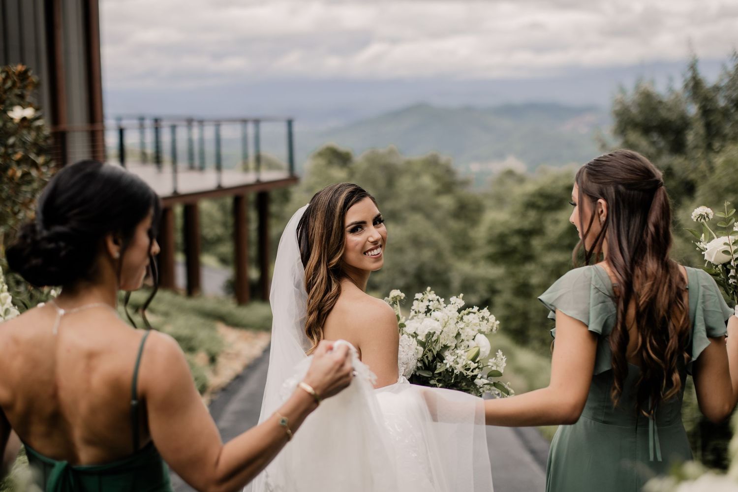 Three women in formal attire gather outdoors with a scenic mountain backdrop.